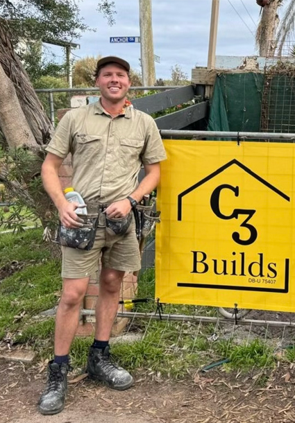 A man in work shirt and shorts standing outdoors next to a yellow sign that says 'C3 Builds'.