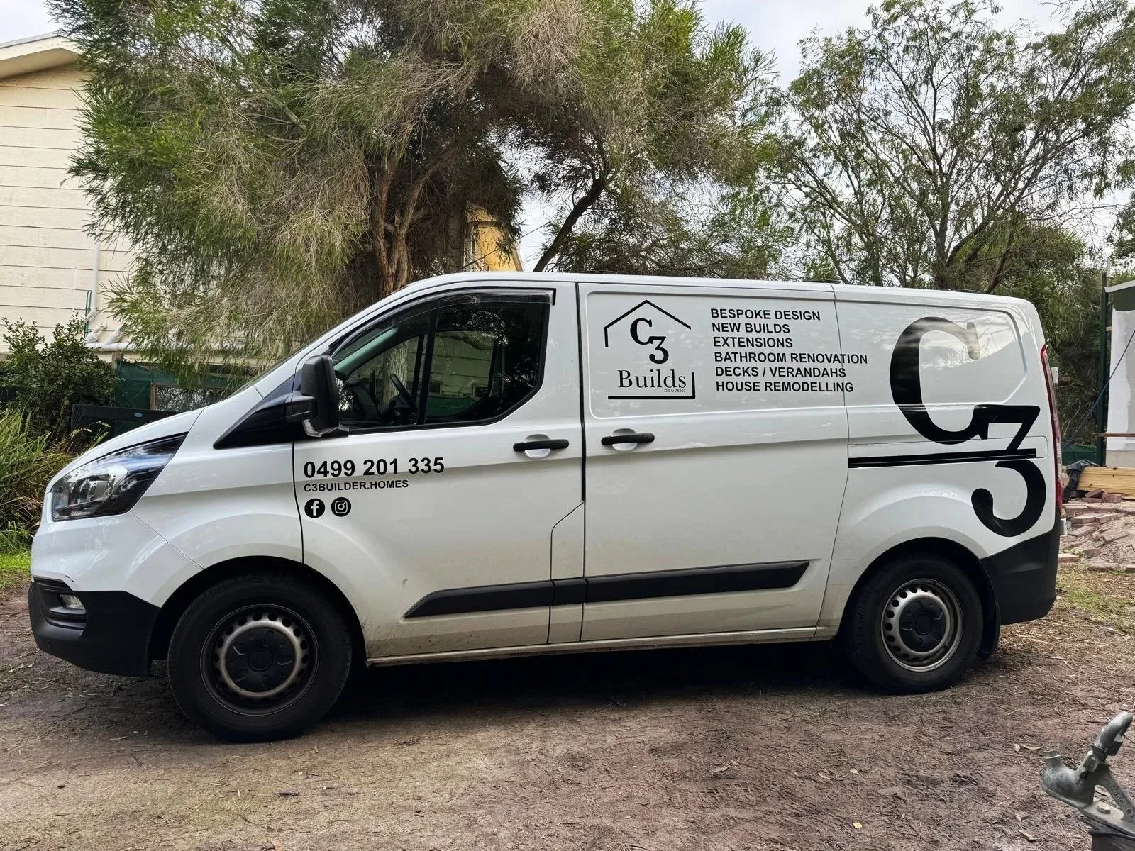 White company van with black lettering advertising C3 Builds, a home renovation business, parked on an unpaved surface with trees and a building in the background.