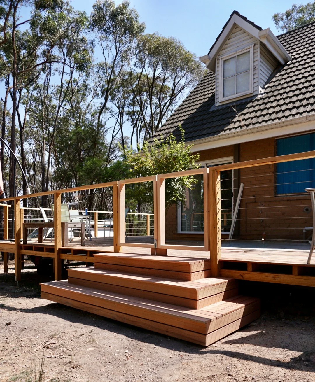 A newly built wooden deck with stairs attached to a brick house with a dormer window, with outdoor chairs and trees in the background.