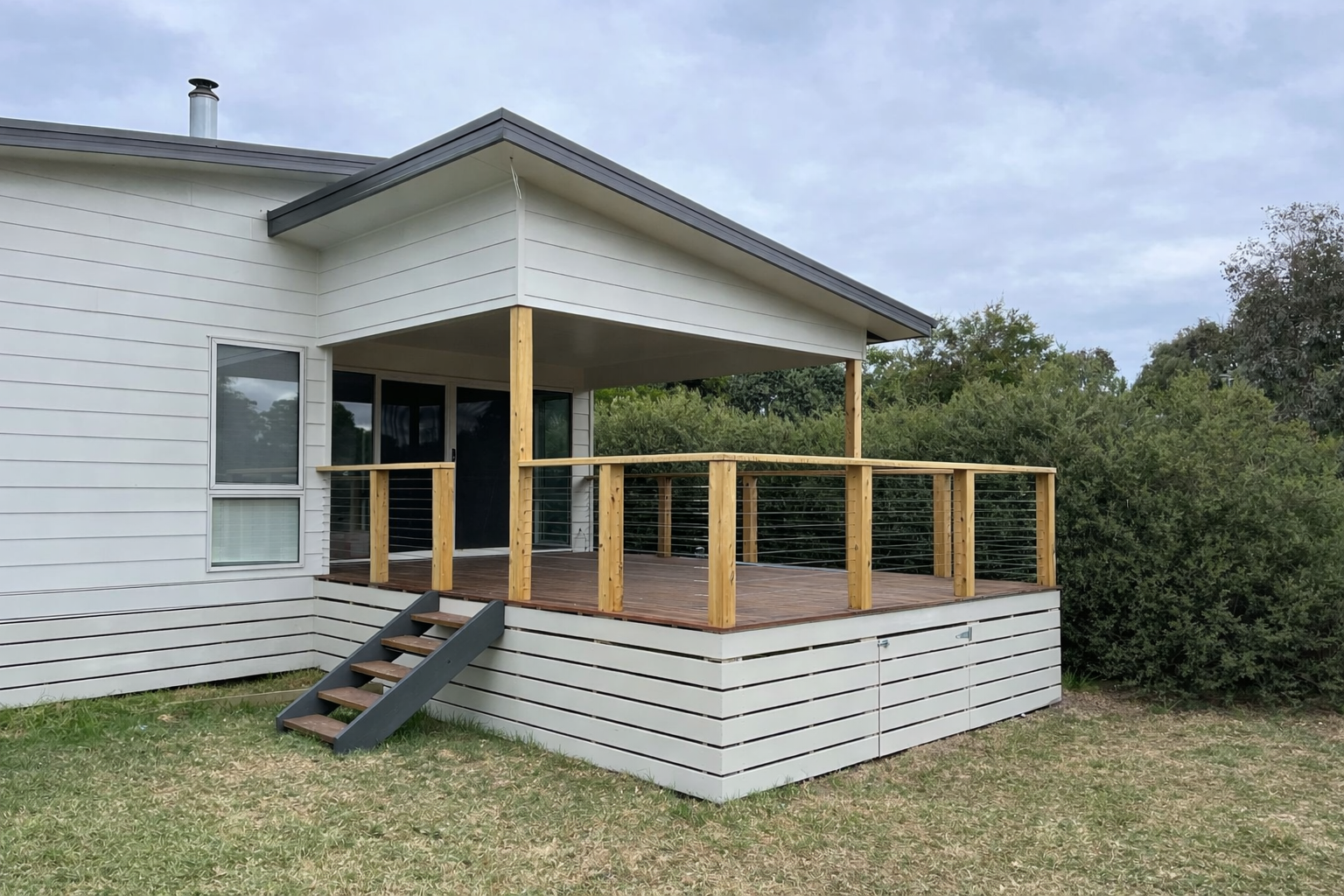A newly built wooden deck attached to a white house with sliding glass doors, featuring a small staircase leading to the grassy yard, and surrounded by trees and overcast sky.