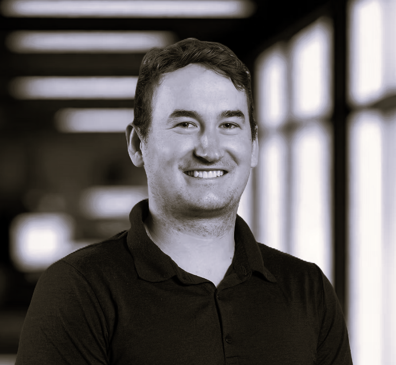 A young man with short dark hair and a friendly smile, wearing a black polo shirt, standing indoors with large windows in the background