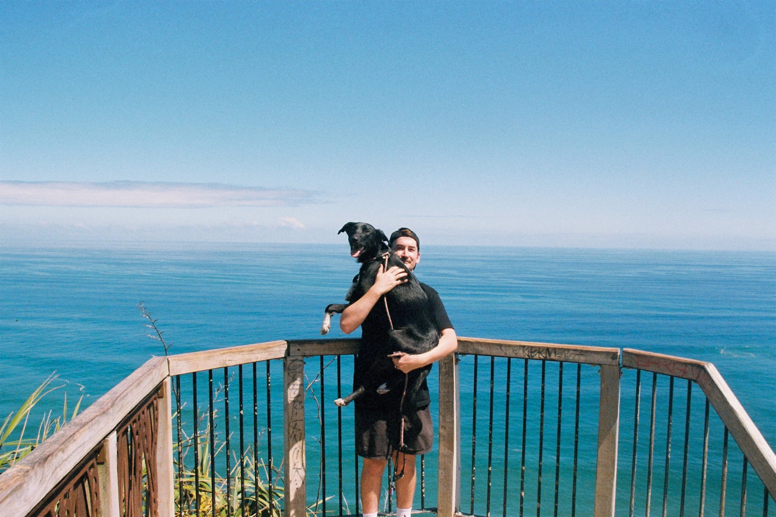 Picture of Corbin Anderson, KiwiSaver Adviser for Voyager Financial. Holding black dog overlooking the sea