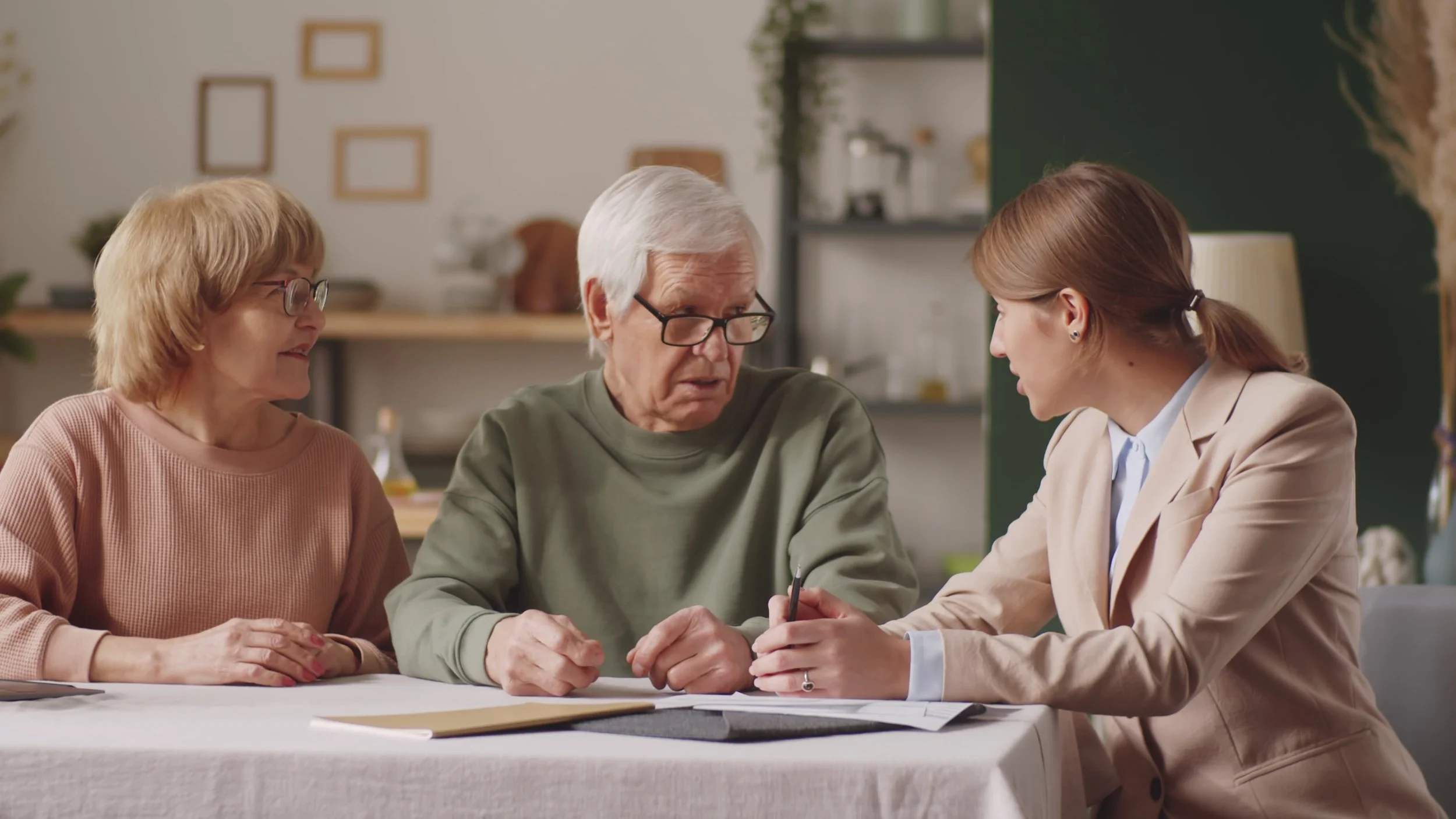 Four people gather around a table, engaged in a serious discussion. An elderly man with white hair and glasses sits in the center, listening intently to a woman with brown hair in a ponytail, wearing a beige blazer, who is speaking to him. An older woman with short brown hair and glasses sits to the left, looking at the elderly man. The background shows a cozy indoor setting with shelves and decorations.