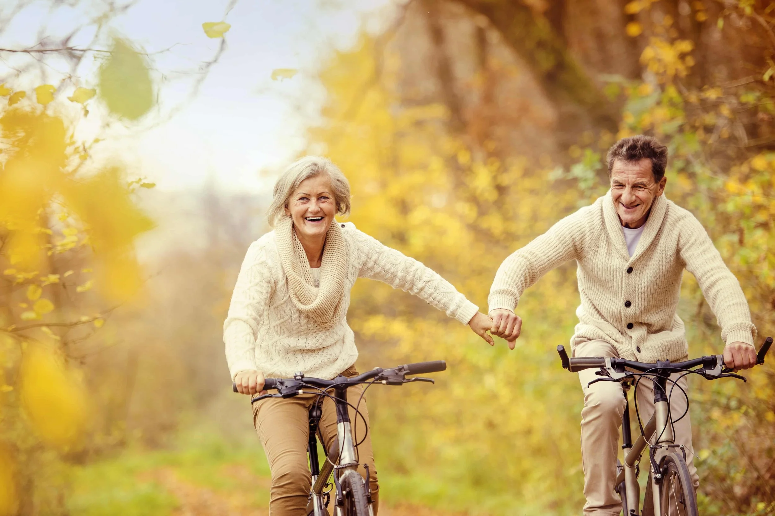 An elderly couple riding bikes together on a fall day in an outdoor setting with yellow and orange leaves.