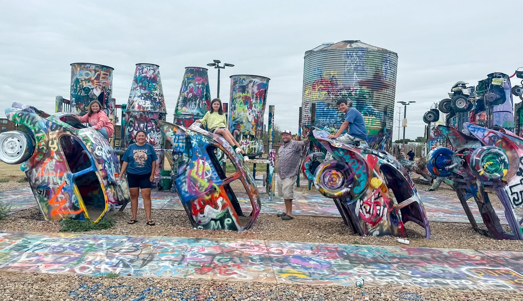 People posing with graffitied sculptures of cars and cylinder shapes in an outdoor art installation.