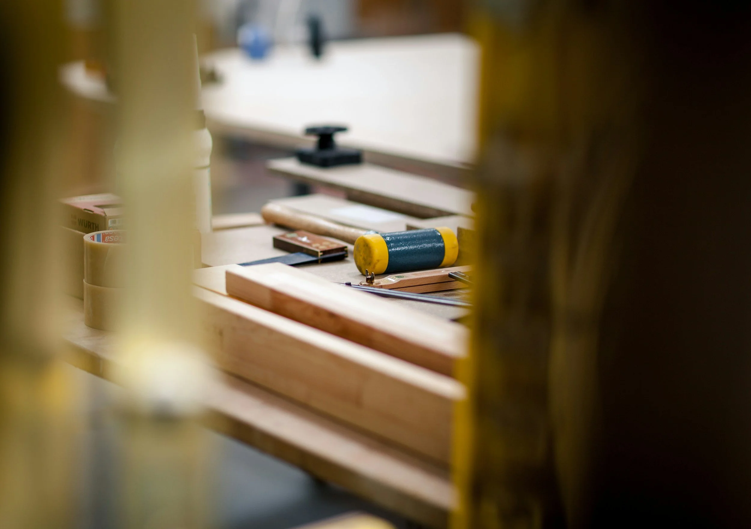 Tools and supplies on a sawmill workbench, including a yellow and blue cylindrical roller and various woodworking clamps, seen through a blurred foreground of yellow workpieces.