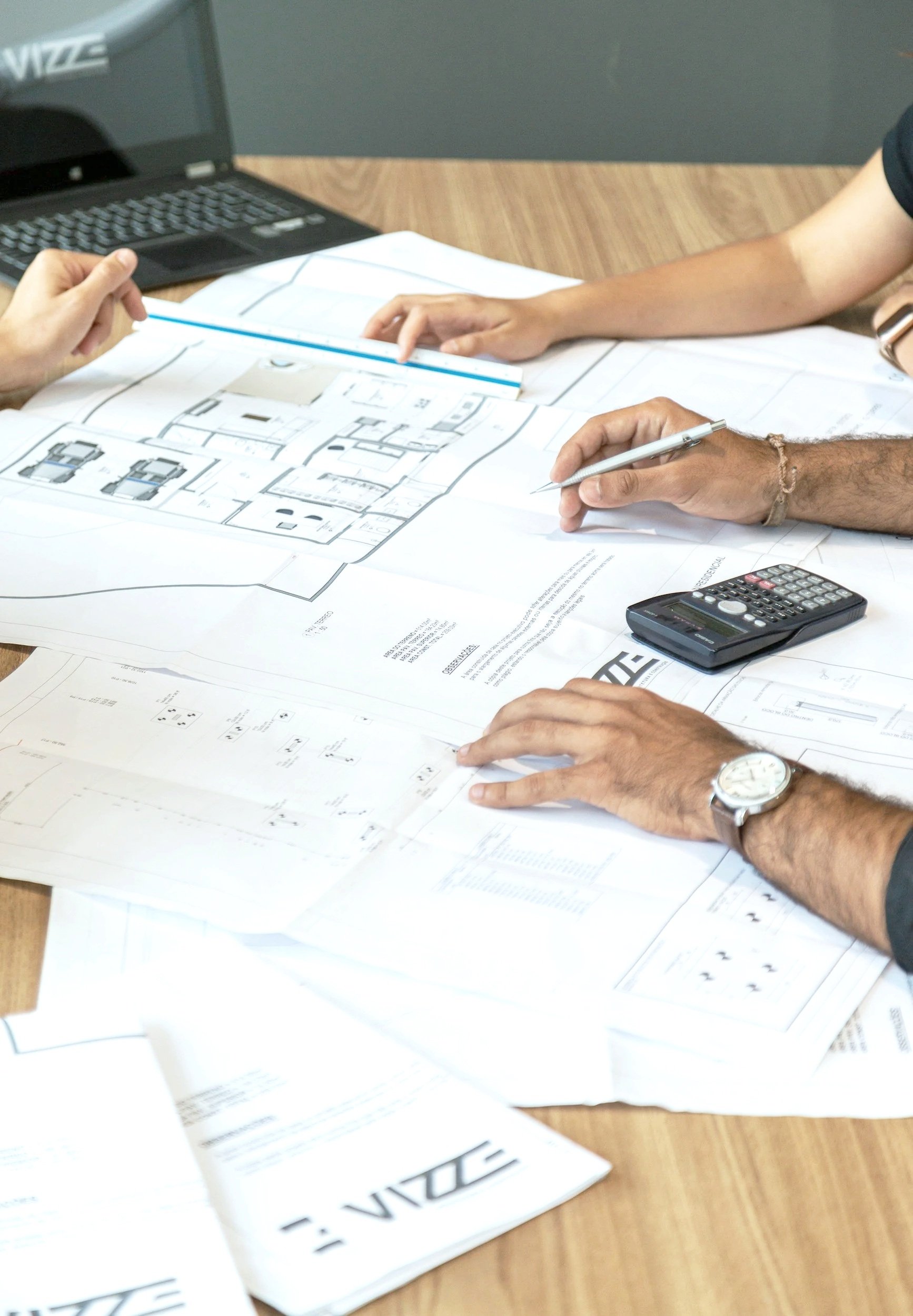 People examining architectural blueprints and plans on a desk with a laptop, calculator, and papers.