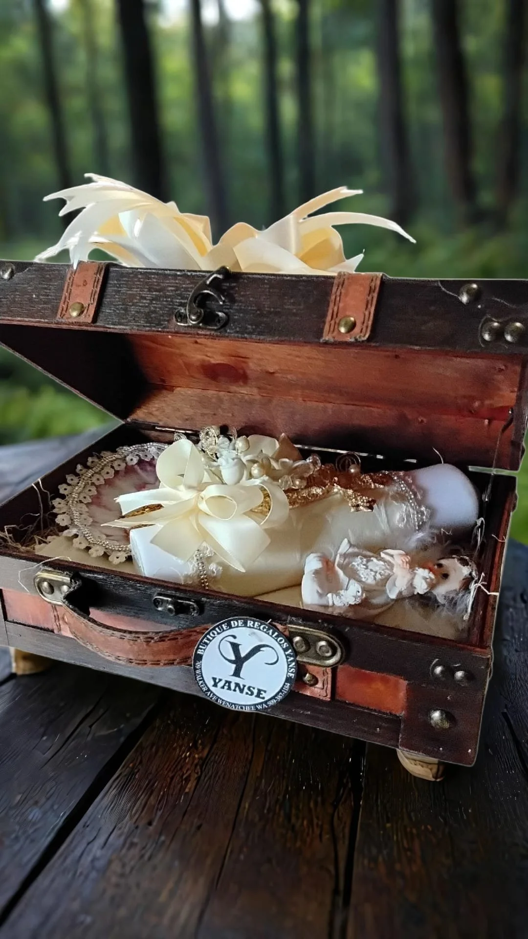 Caja de madera abierta con accesorios blancos y perlas dentro, en un fondo de bosque con árboles y hojas verdes.