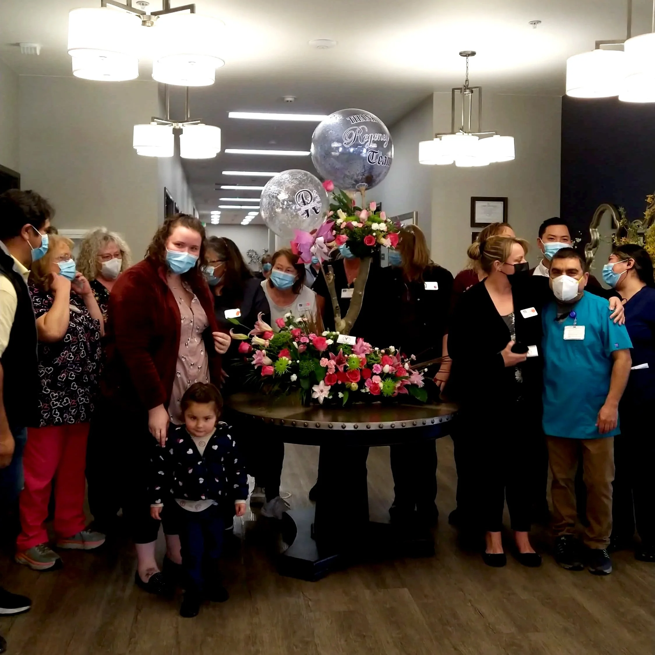 Grupo de personas en una reunión o evento, con máscaras, rodeando una mesa con arreglos florales y globos, en un espacio cerrado con iluminación moderna.