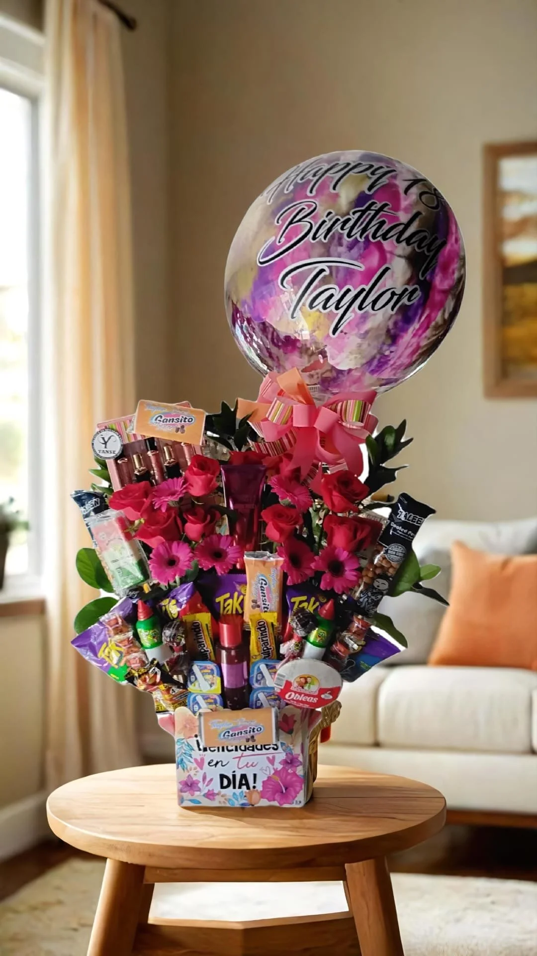 Arreglo de flores y dulces con globo que dice 'Feliz cumpleaños Taylor' sobre mesa de madera en sala.