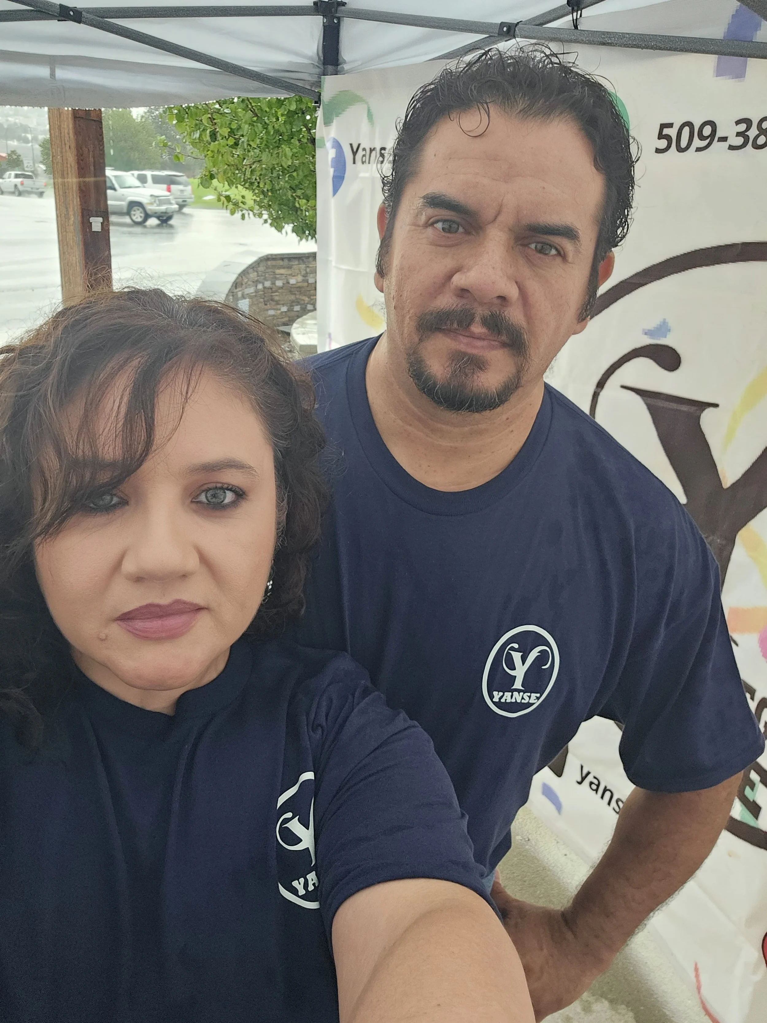 Dos personas con camisetas azules con logotipo de Yanse toman una selfie en un puesto de exhibición al aire libre, en un día lluvioso.