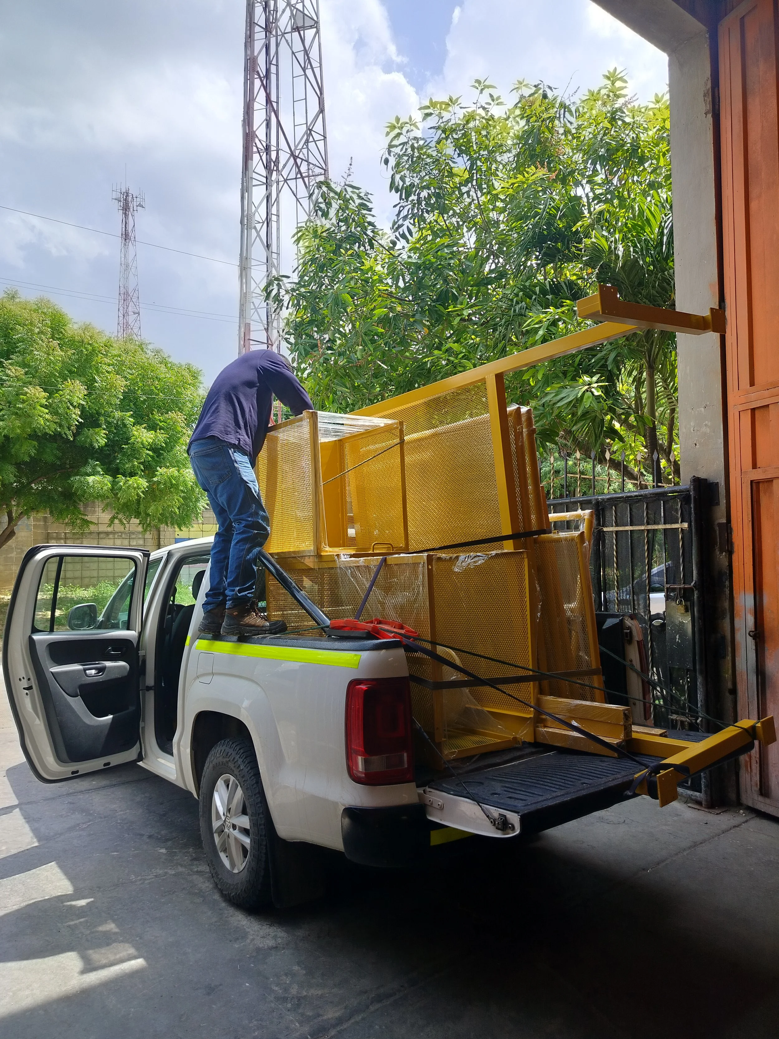 Un hombre colocando equipo de telecomunicaciones en la caja de una camioneta blanca, con dos torres de transmisión de fondo y árboles verdes.