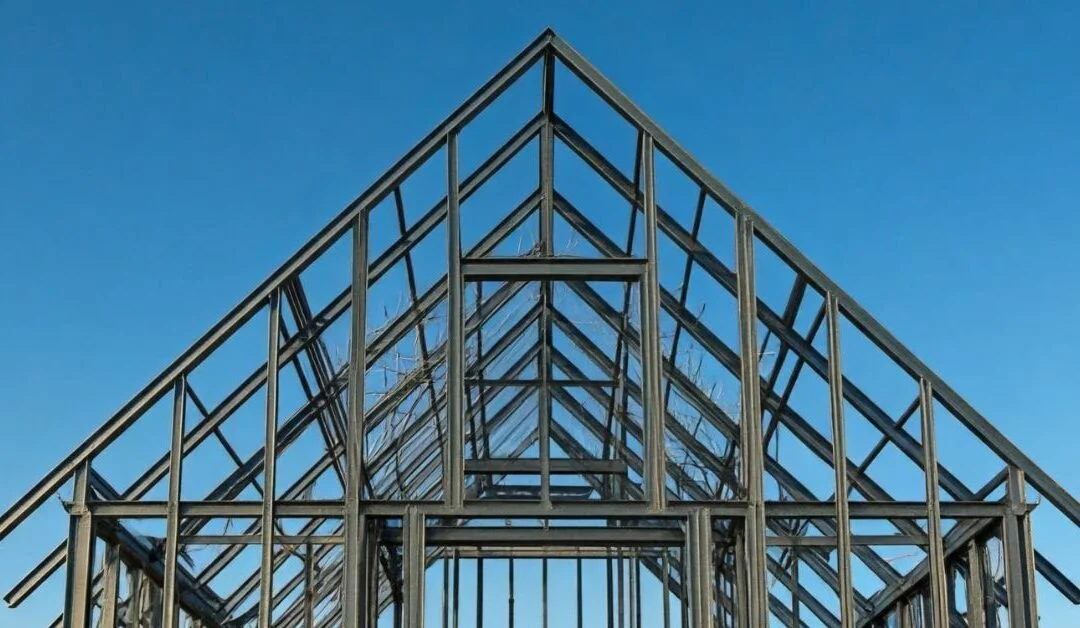 Estructura metálica de un invernadero con marco de acero y techo de vidrio o plástico transparente, bajo un cielo azul.