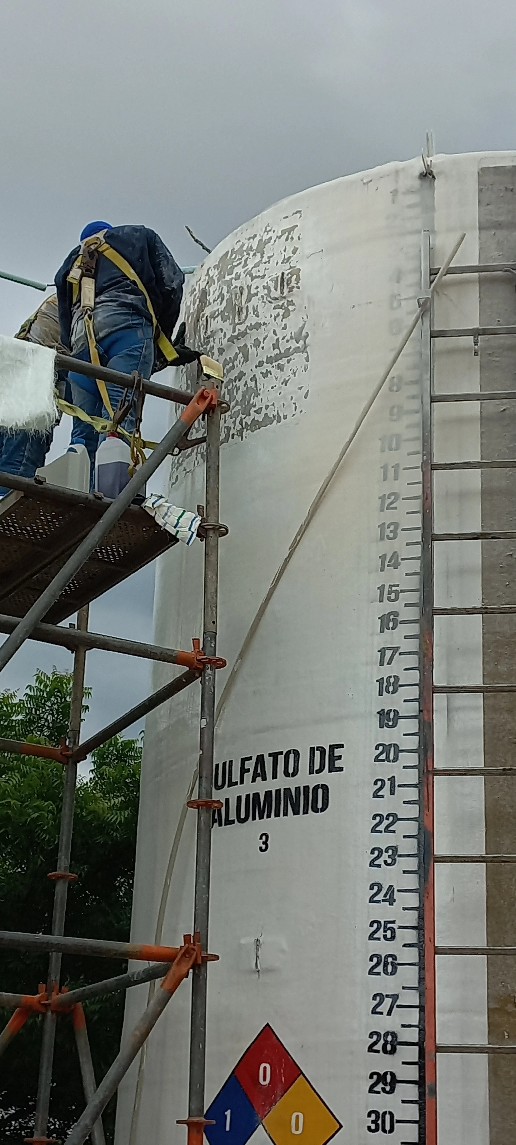 Trabajadores pintando un tanque grande de aluminio con pintura especializada, en un día nublado.