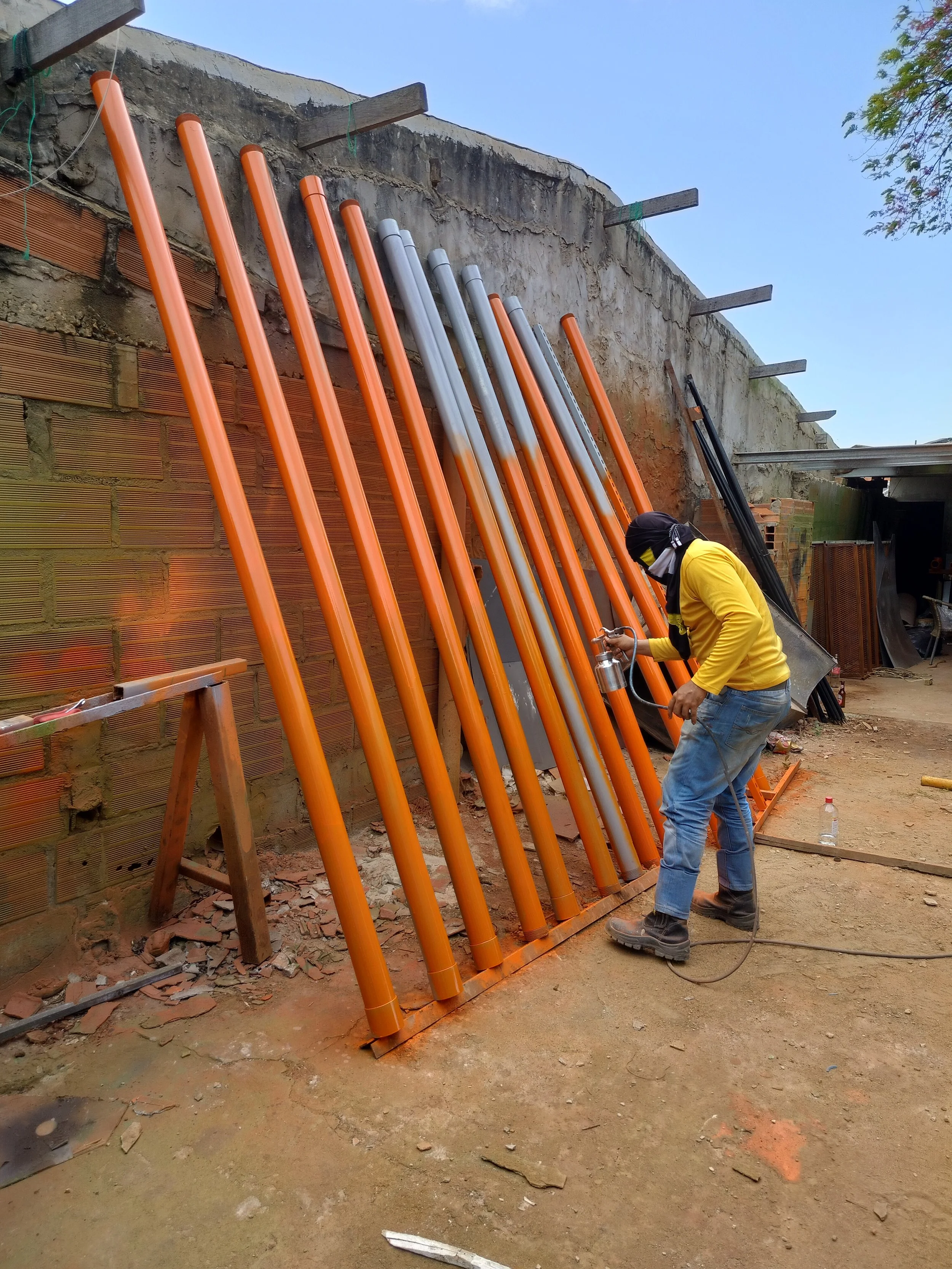 Un trabajador en uniforme amarillo con máscara, utilizando una herramienta de soldar, trabaja con tubos de electricidad naranja y gris en una obra de construcción. La pared de ladrillos y la estructura de madera se aprecian en el fondo.