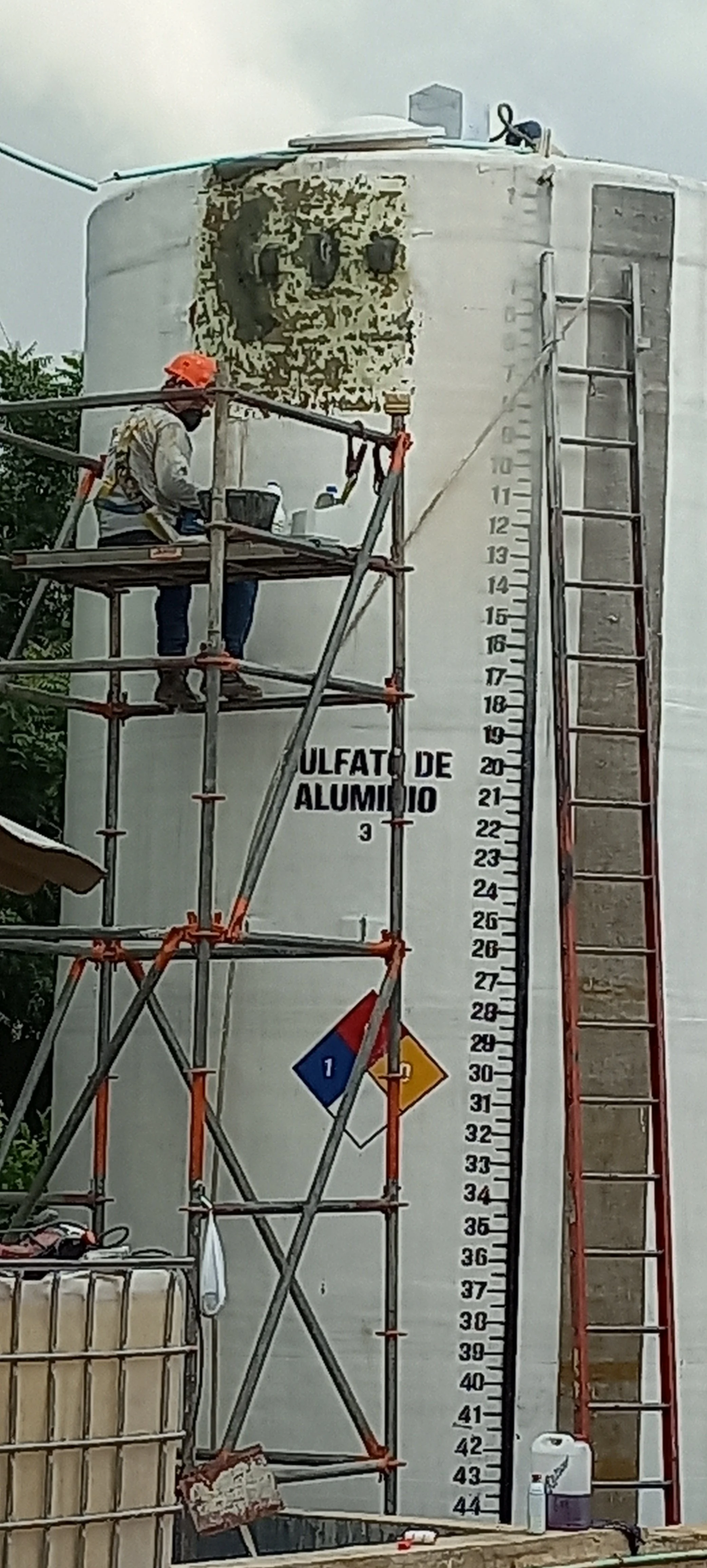 Trabajador en andamios pintando un tanque de aluminio, que tiene una escala de medición y el texto 'OLFACTO DE ALUMINIO' en la superficie.