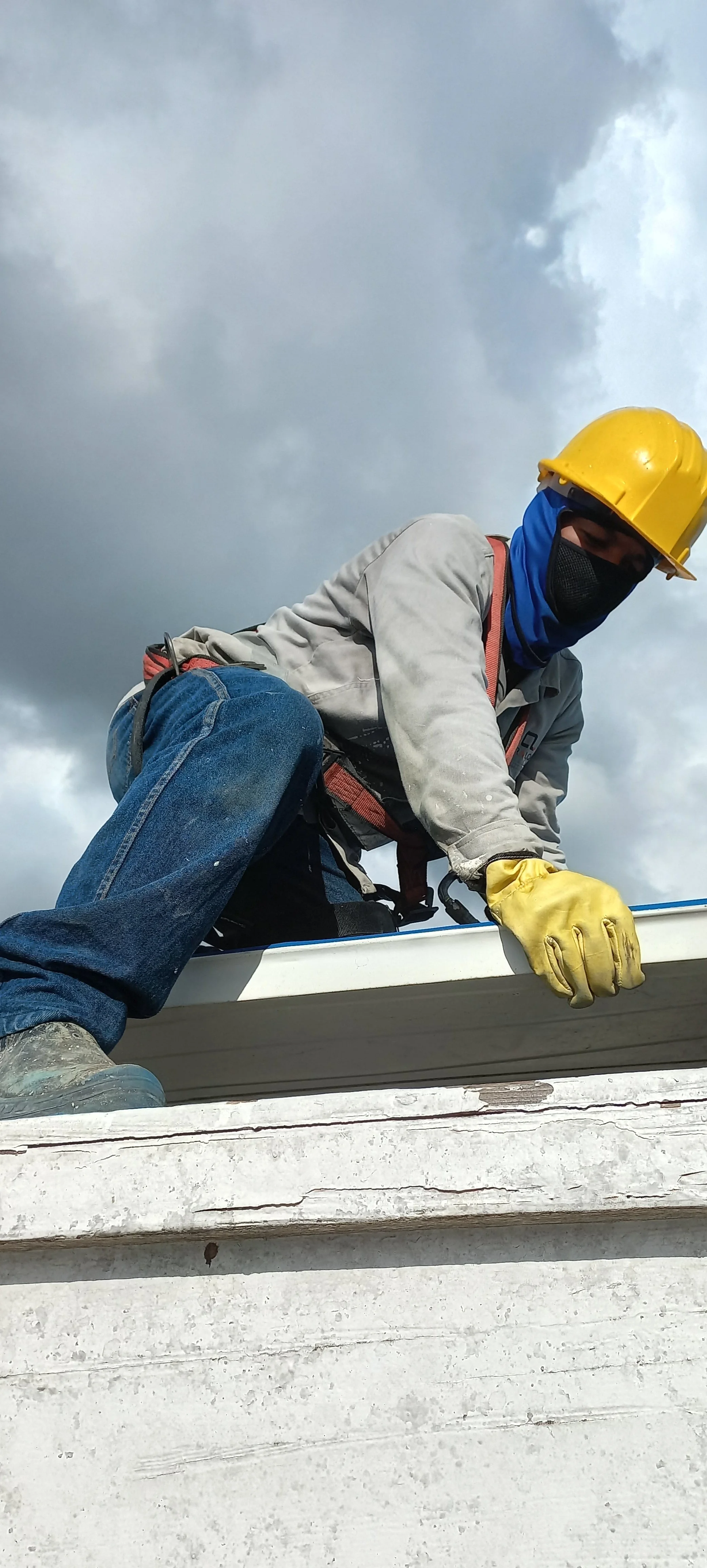 Trabajador usando casco amarillo, guantes amarillos, máscara negra, chaleco de seguridad, y ropa de trabajo en un techo, bajo cielo nublado.