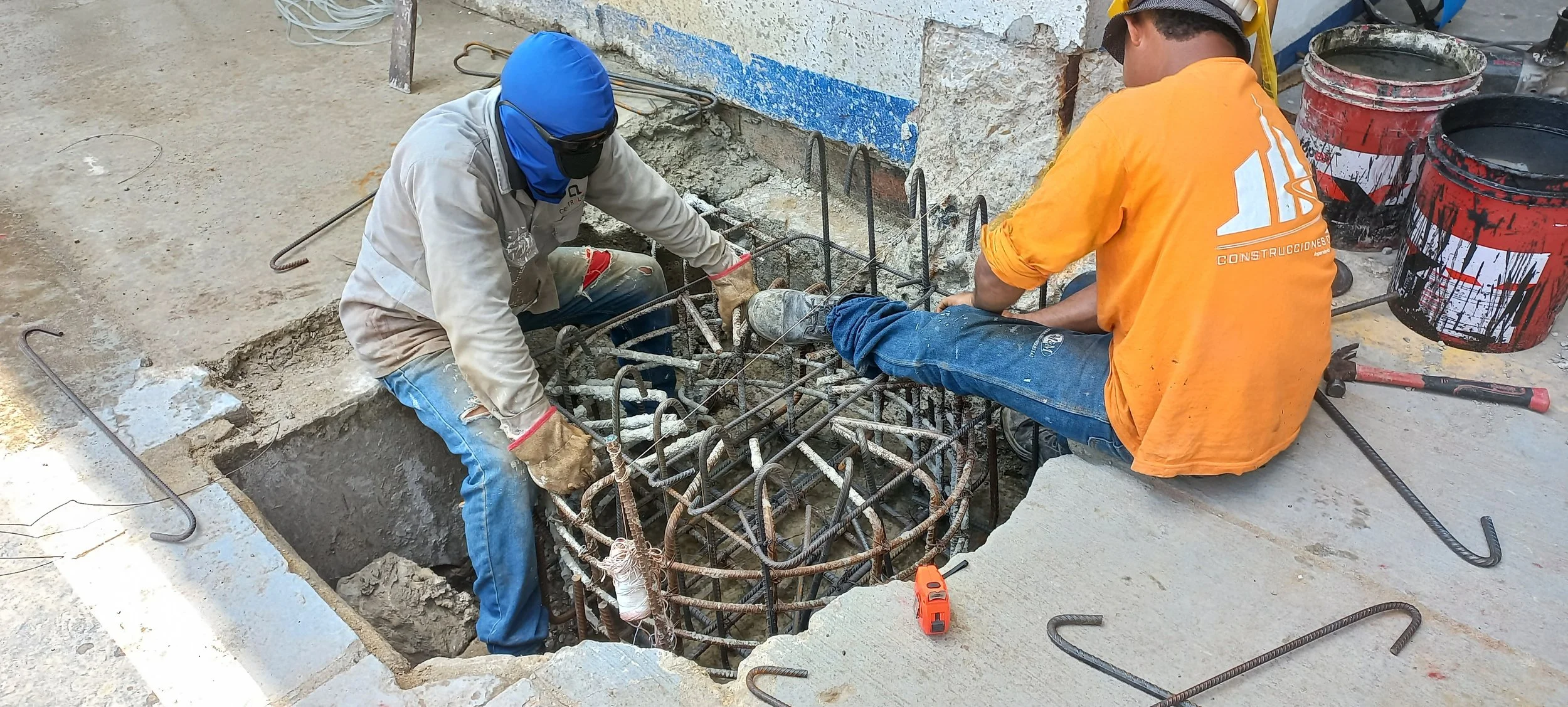 Dos trabajadores de construcción colocando acero de refuerzo en una excavación, uno con casco y el otro con cubierta facial y guantes, en un trabajo de cimentación.