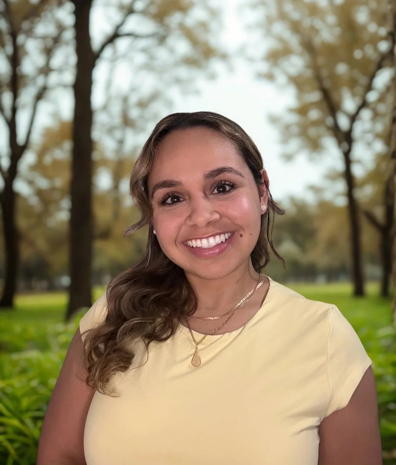 A woman with wavy brown hair smiling outdoors in a park with trees and grass in the background, wearing a yellow top and layered necklaces.