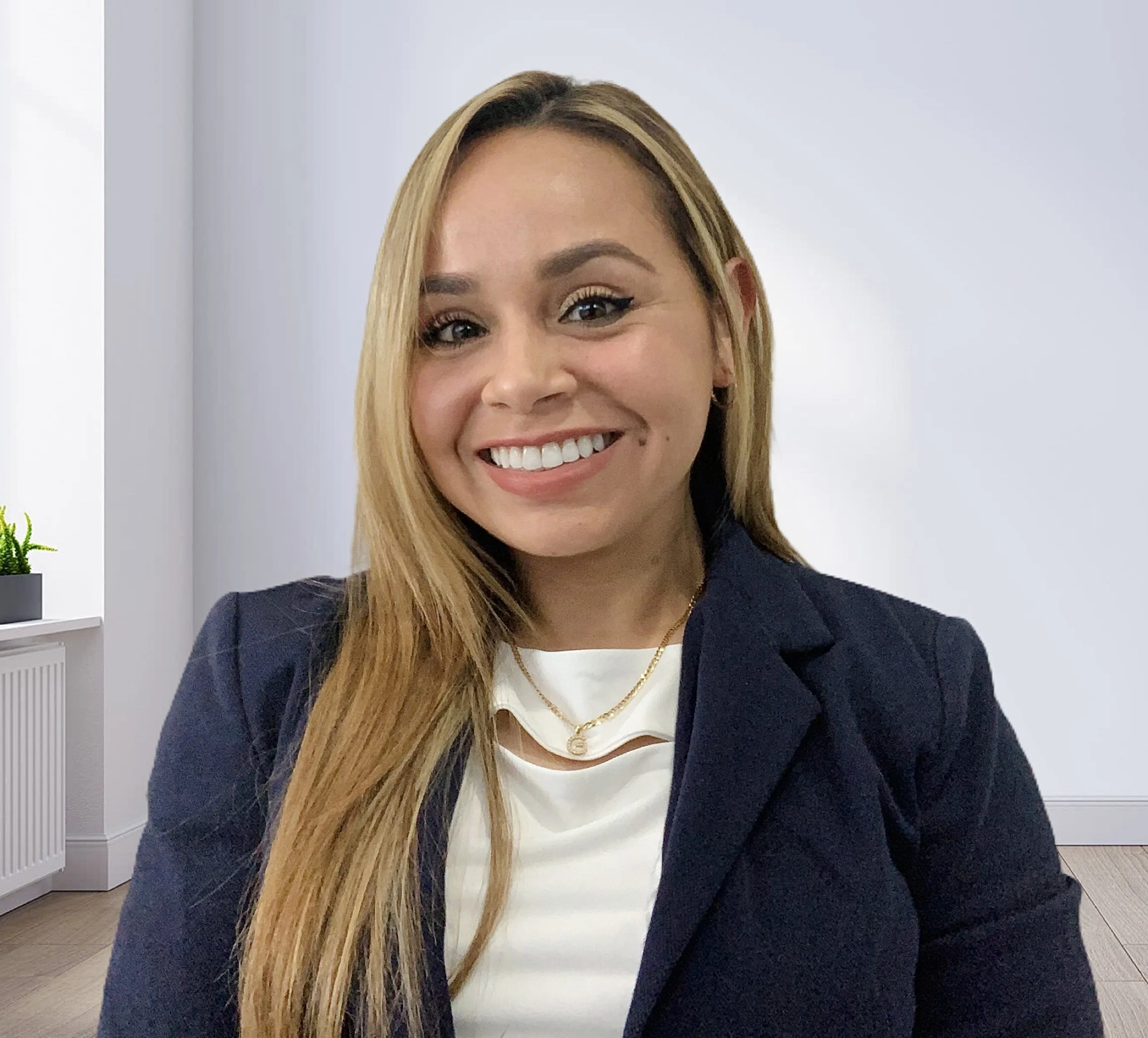 A woman with long blonde hair smiling, dressed in a navy blazer and white top, wearing a gold necklace, in a bright office setting.