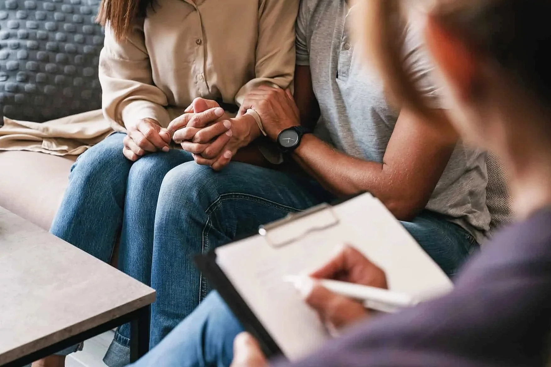 Two people holding hands during a counseling session, with a therapist taking notes on a clipboard.