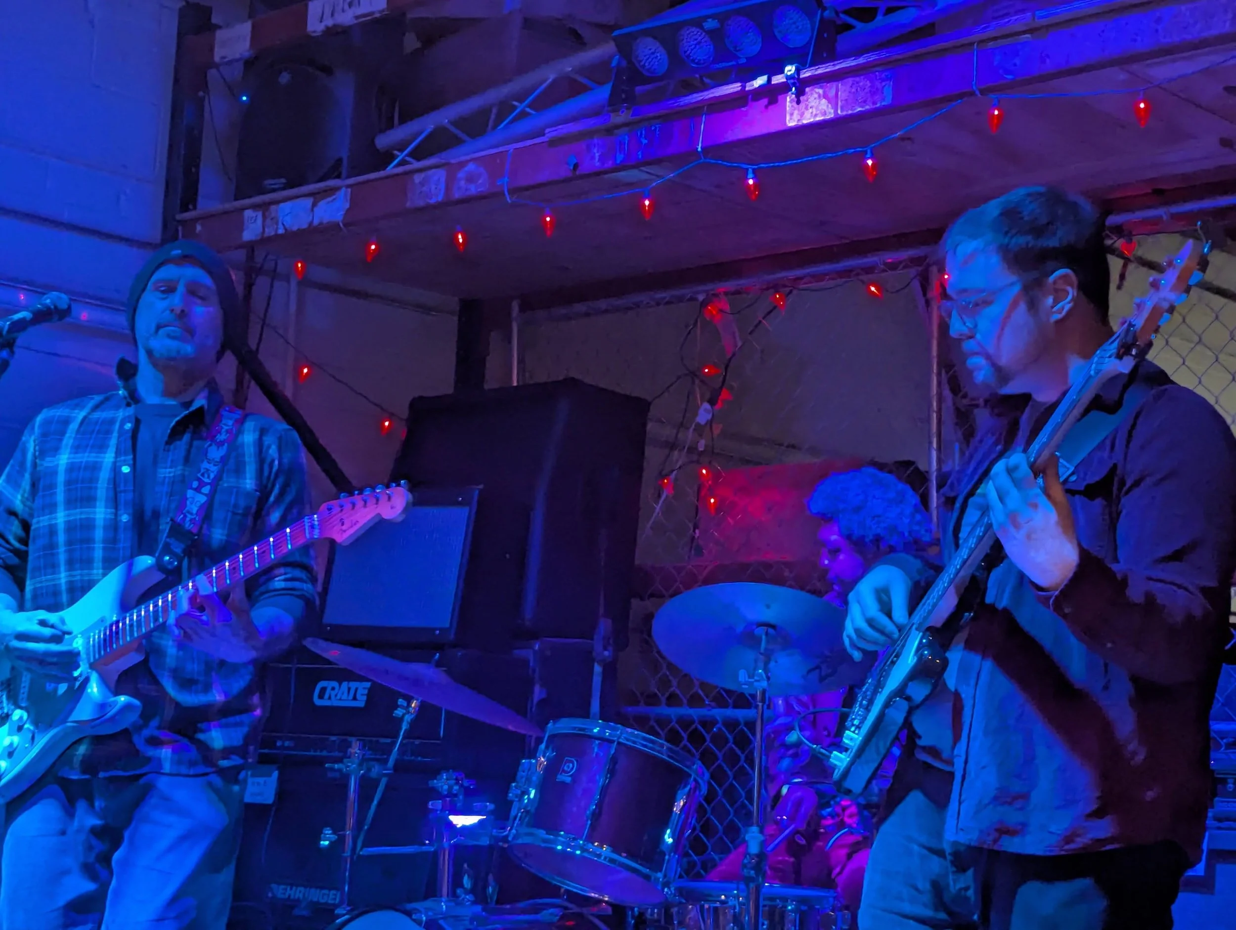 Three musicians perform in a dimly lit venue with purple and blue lighting, one playing an electric guitar, another on drums, and the third on bass guitar, with red string lights hanging above.