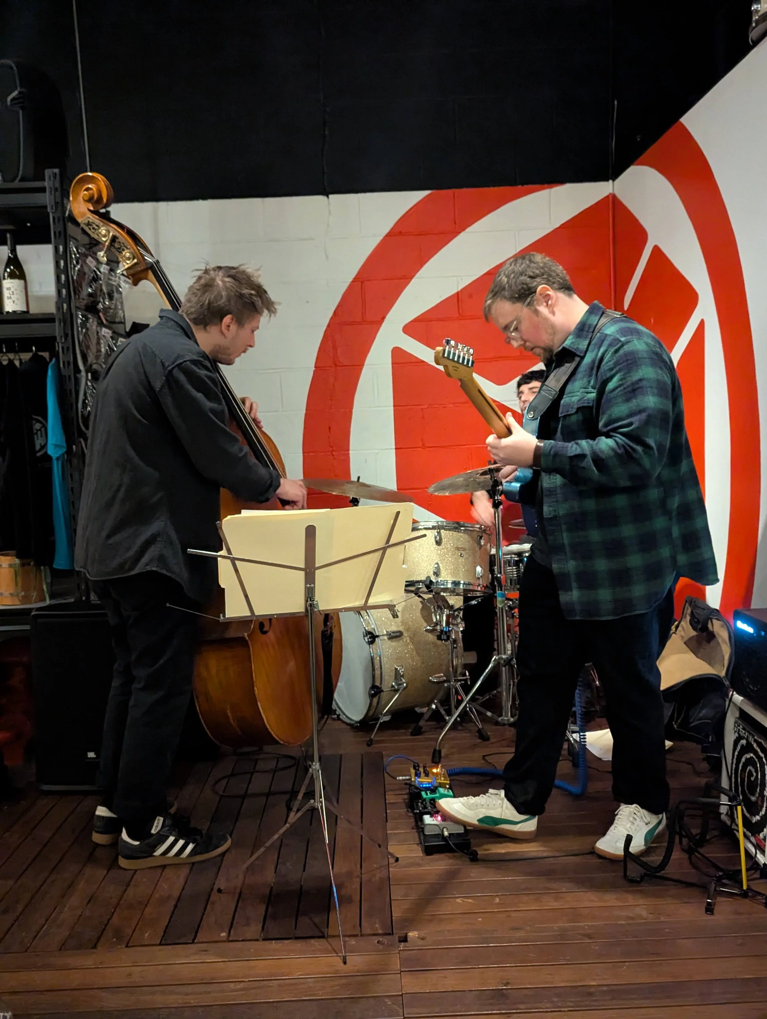 Three musicians playing instruments in a band: a bassist, a guitarist, and a drummer. They are performing in a room with a colorful mural on the wall behind them.