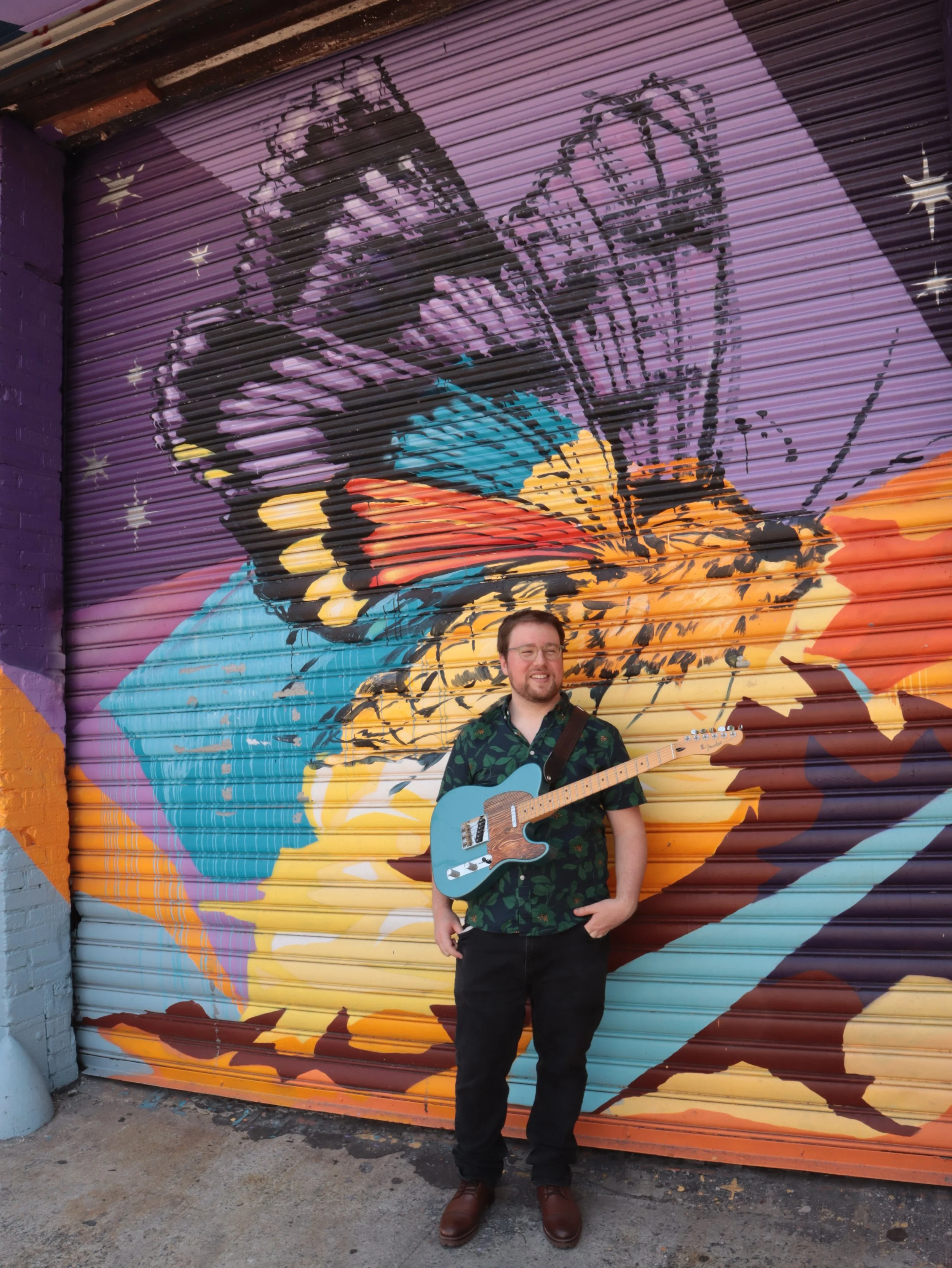 A man with glasses and a beard standing in front of a colorful butterfly mural on a purple garage door, holding a blue electric guitar.