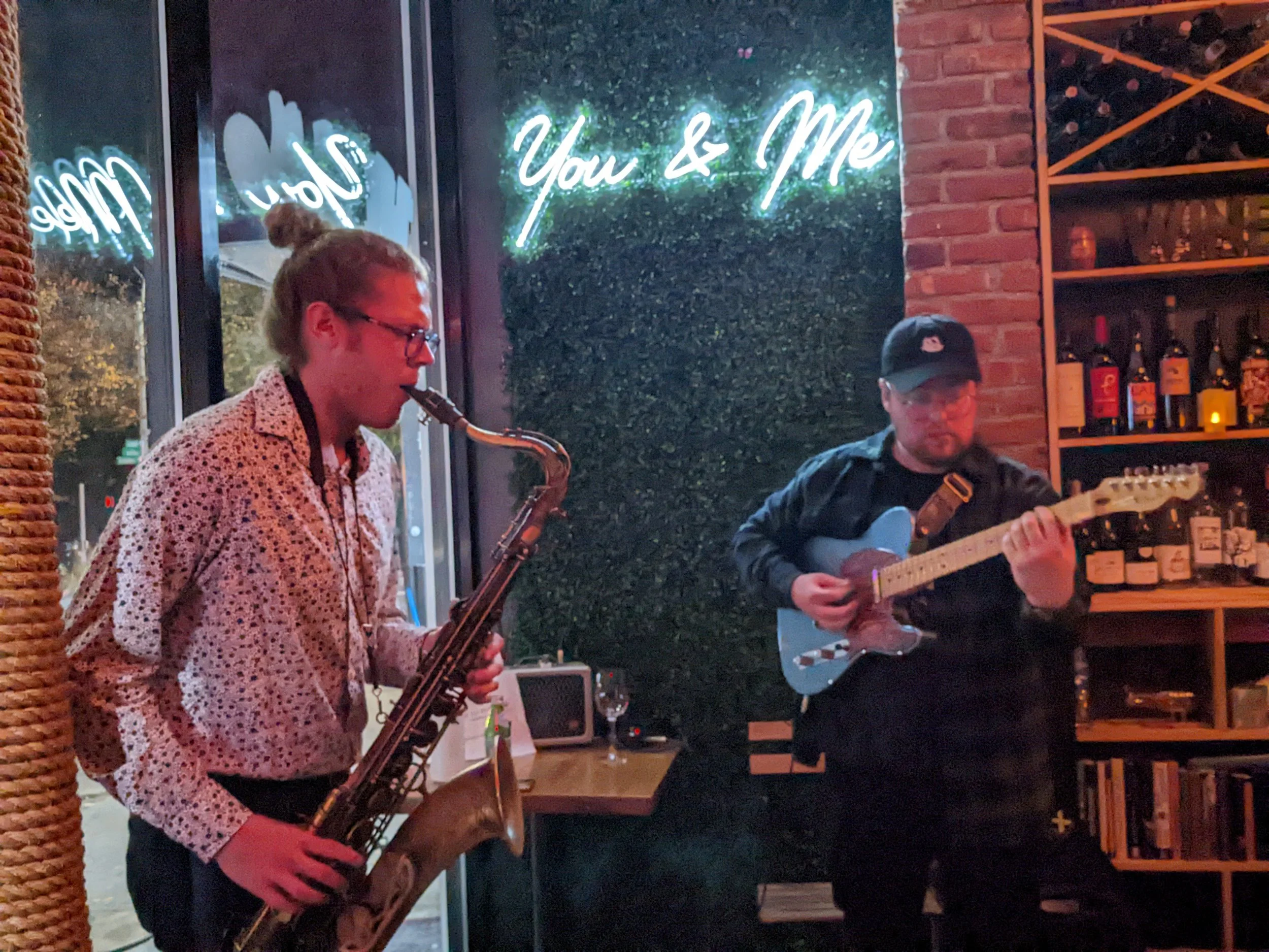 Two musicians playing instruments in a dimly lit restaurant or bar. The person on the left is playing the saxophone, and the person on the right is playing the guitar. There is a neon sign in the background that says 'You & Me' and a bookshelf with bottles and books.