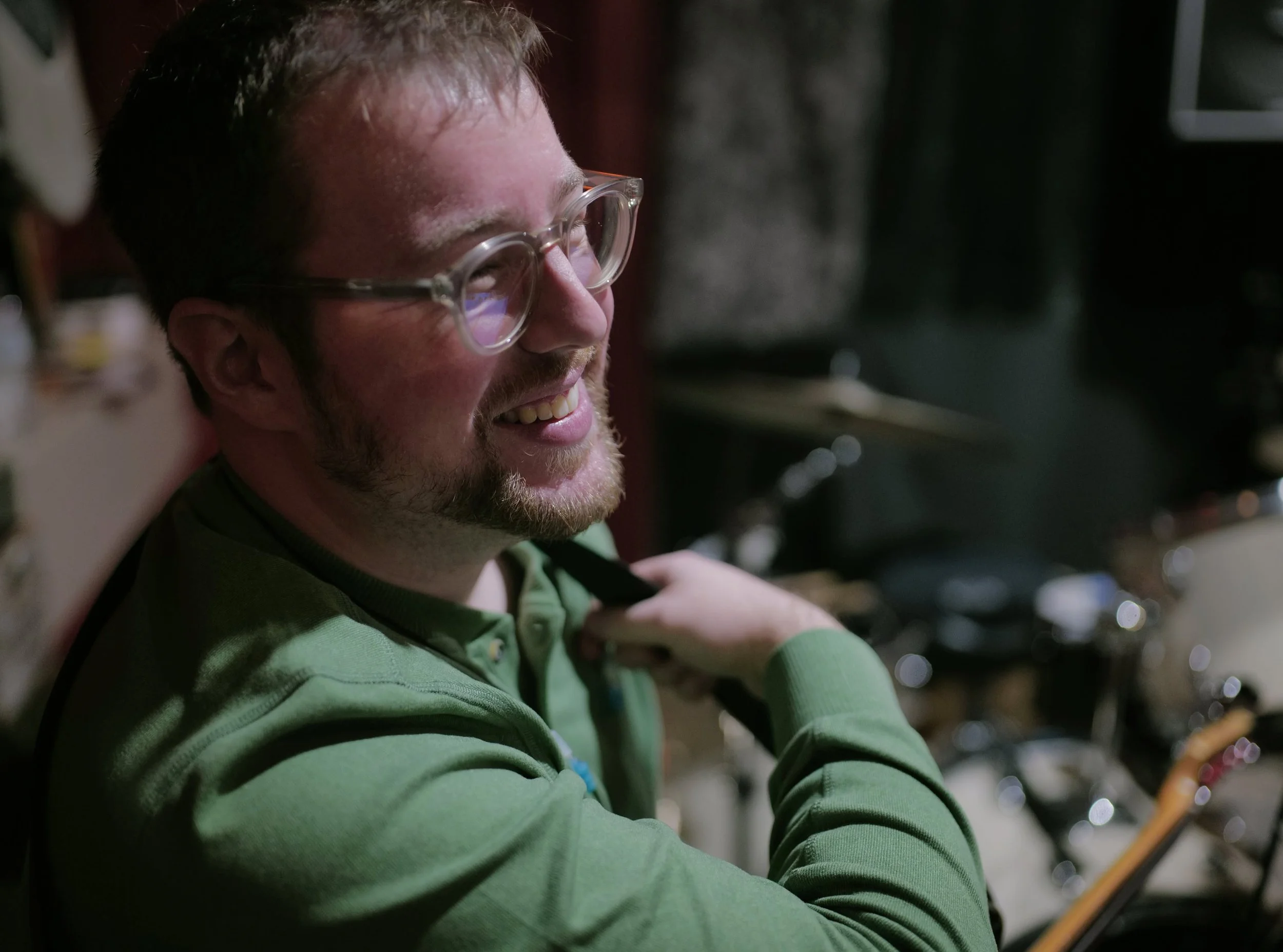 A man with glasses and a beard smiling, wearing a green shirt, sitting in a dimly lit room with musical instruments in the background.