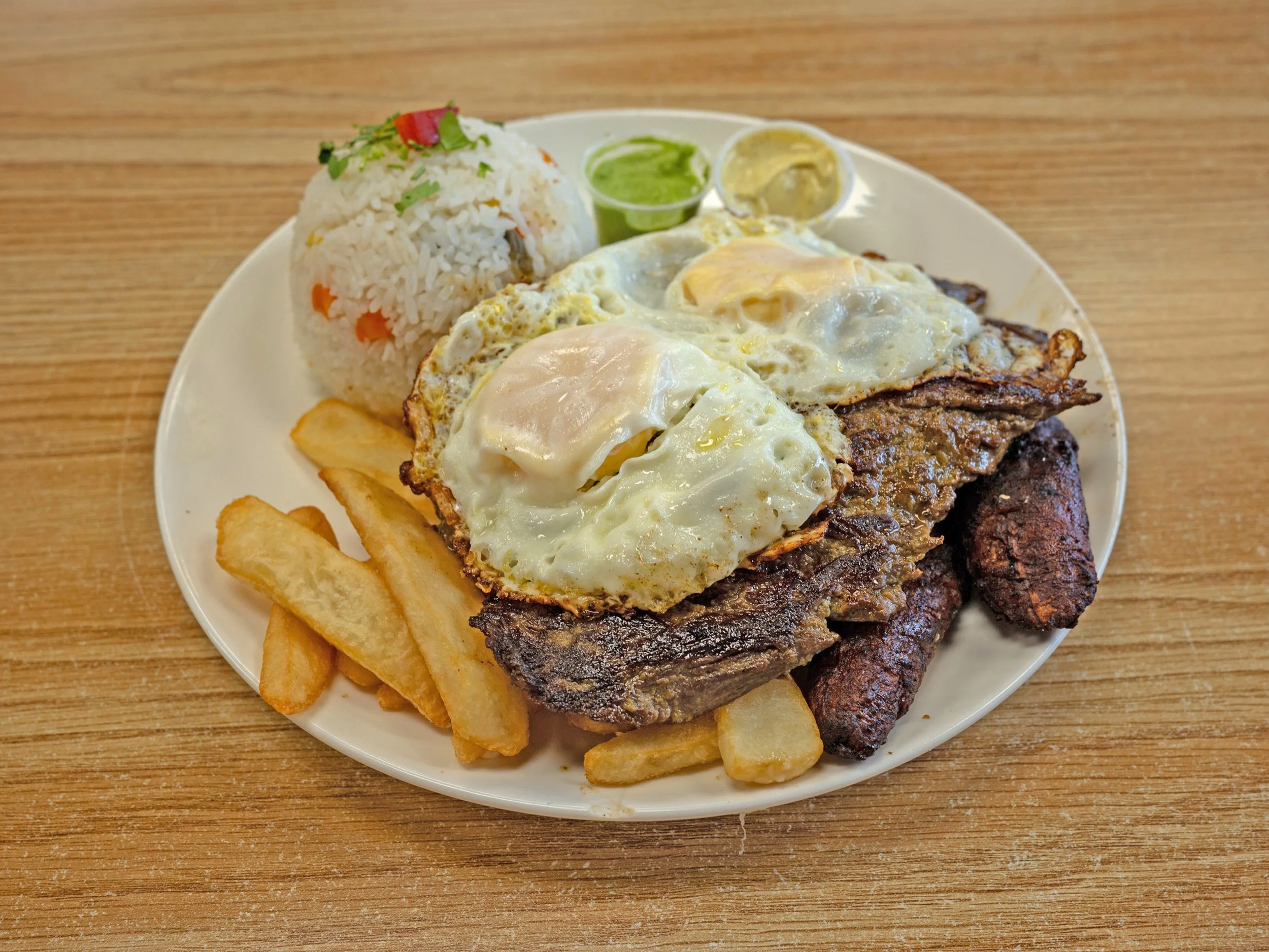 Plate of fried eggs on beef steak, served with rice, French fries, plantains, and side sauces on a wooden table.