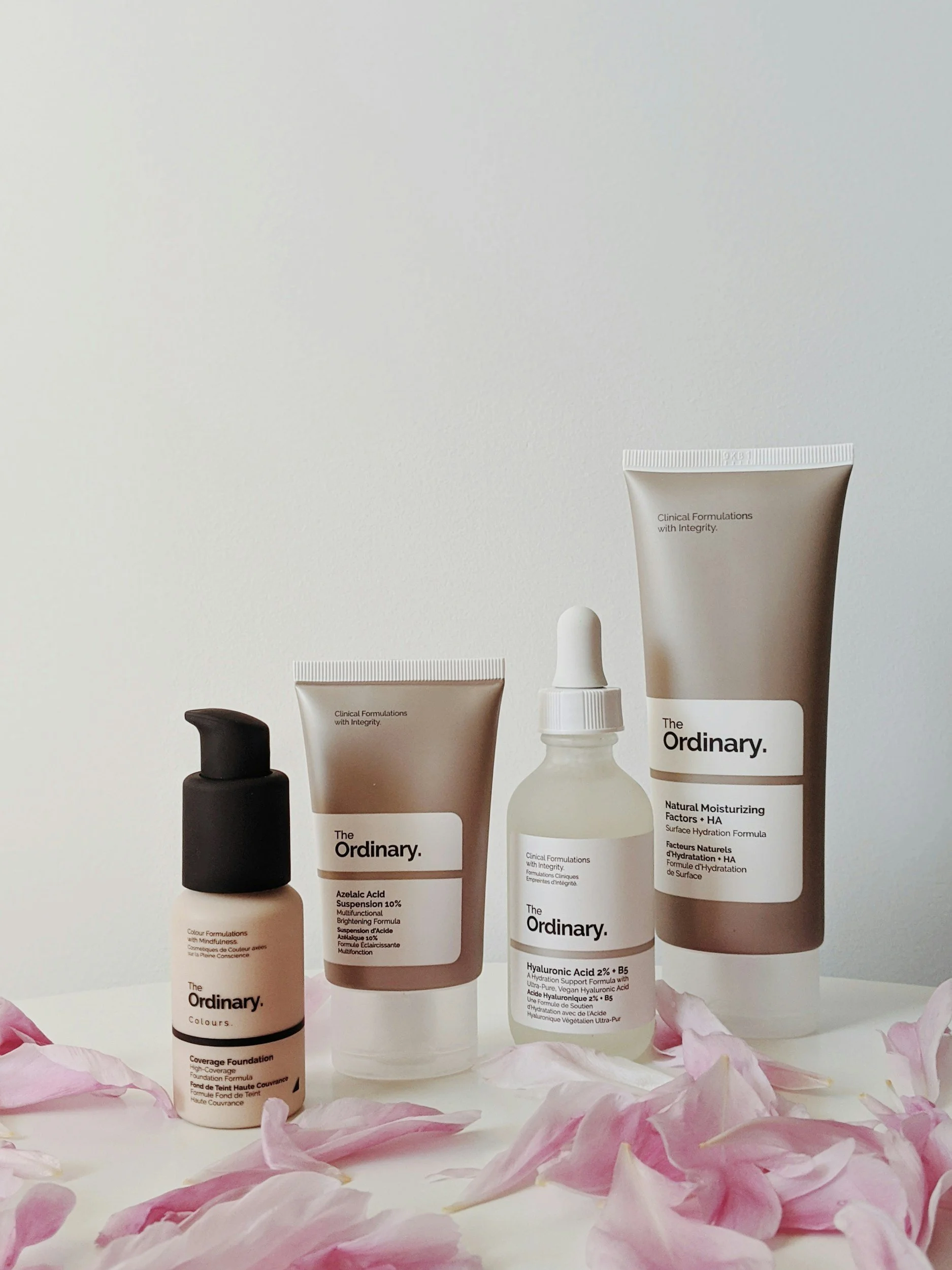 A collection of skincare products from The Ordinary on a white surface surrounded by pink flower petals.