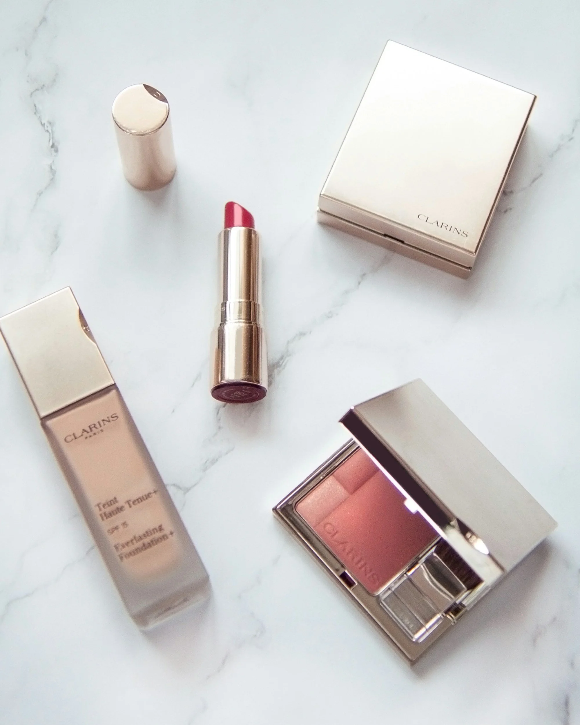Collection of Clarins skincare and makeup products including foundation, lipstick, blush, and powders on a white marble surface.