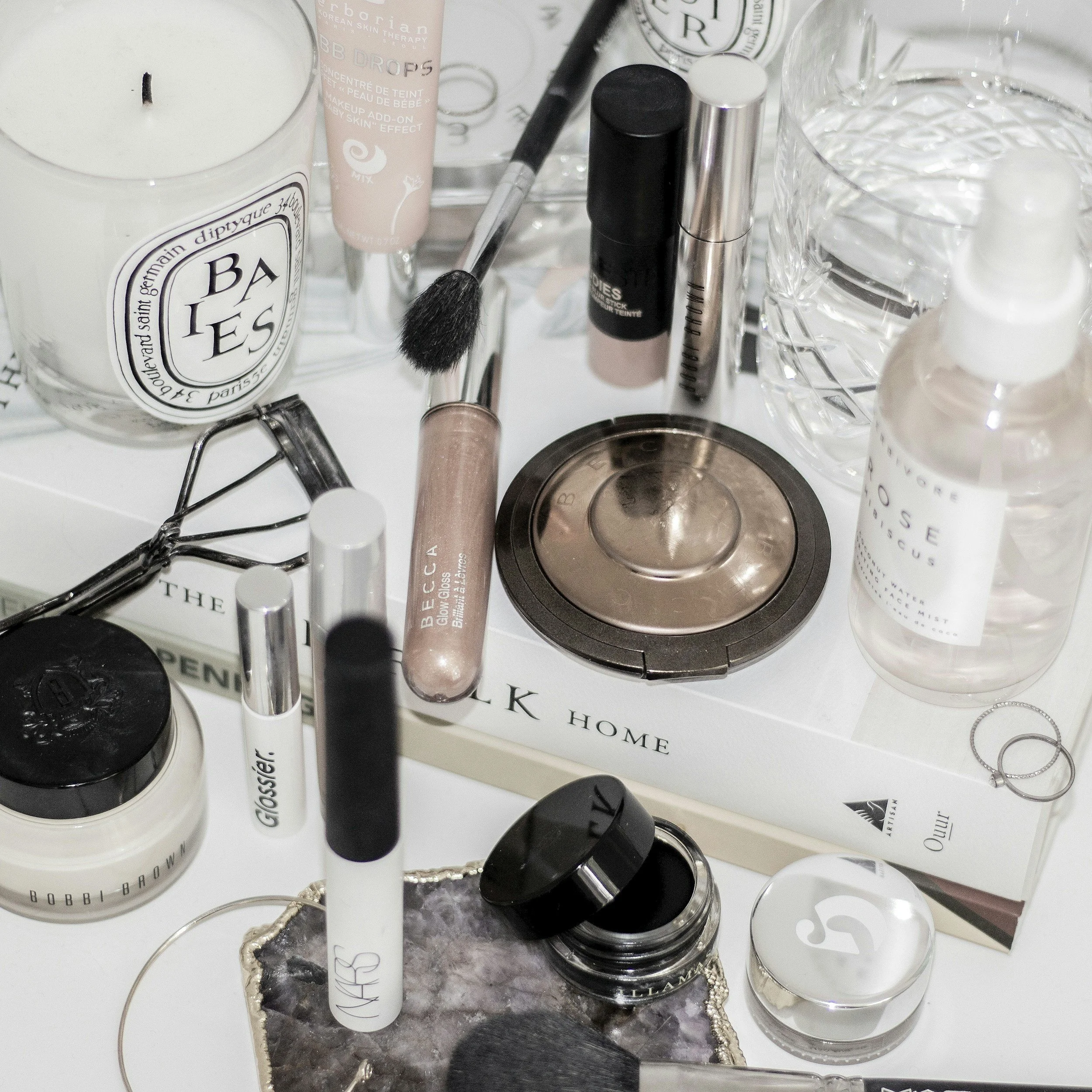 A collection of makeup and skincare products, including a candle, brushes, a highlighter, a setting spray, and various cosmetic containers on a white surface.