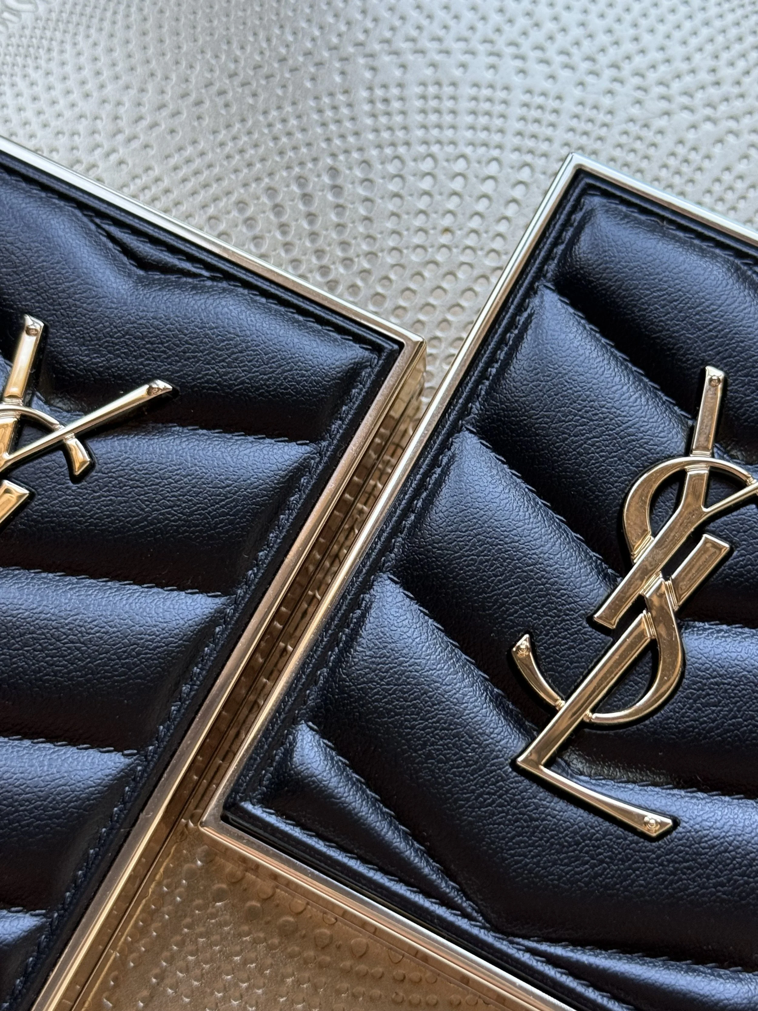 Close-up of two black quilted bags with gold YSL logos and gold frames, placed on a beige textured surface.