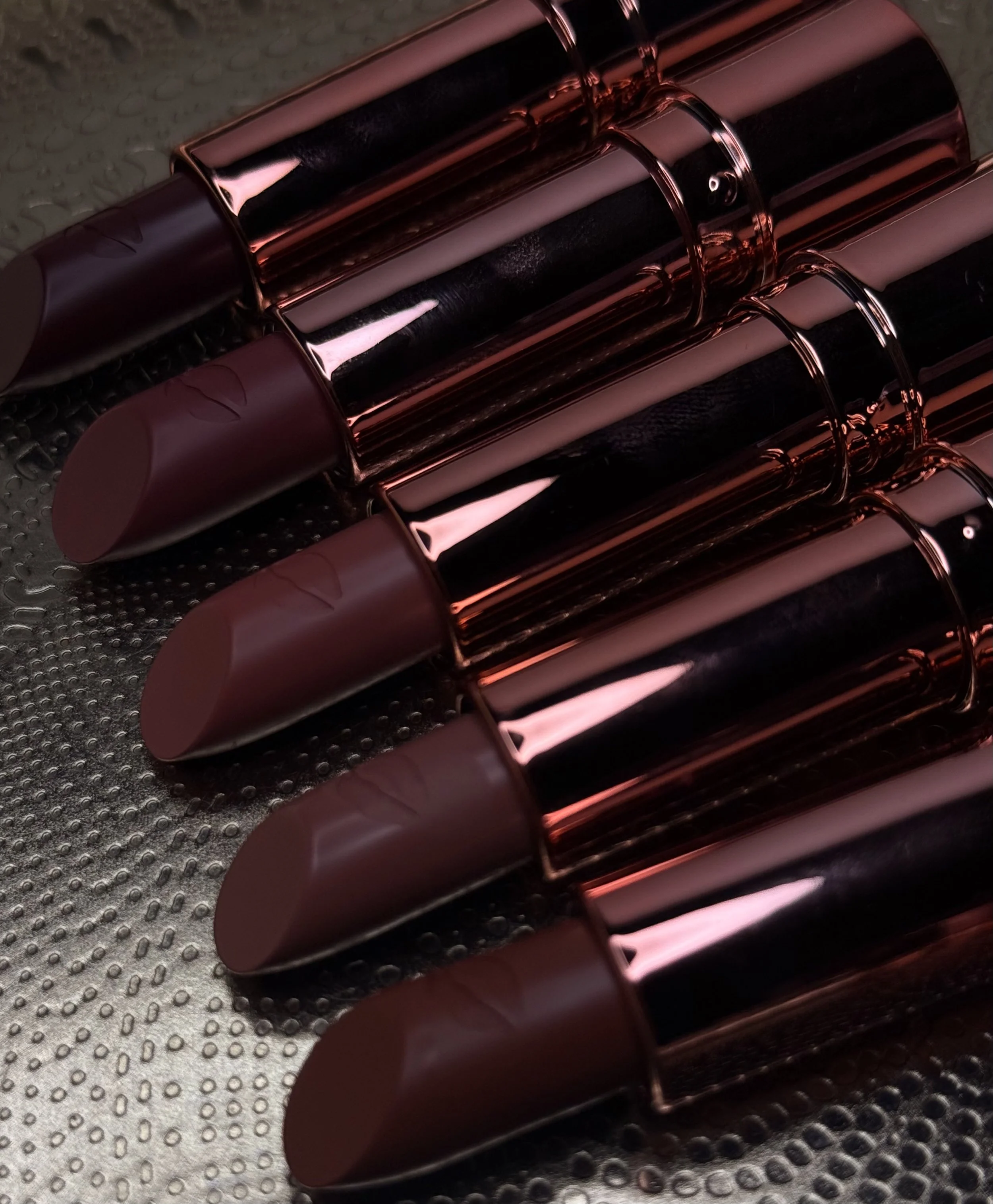 Four matte lipstick tubes with metallic rose gold caps and black bodies arranged diagonally on a textured surface.