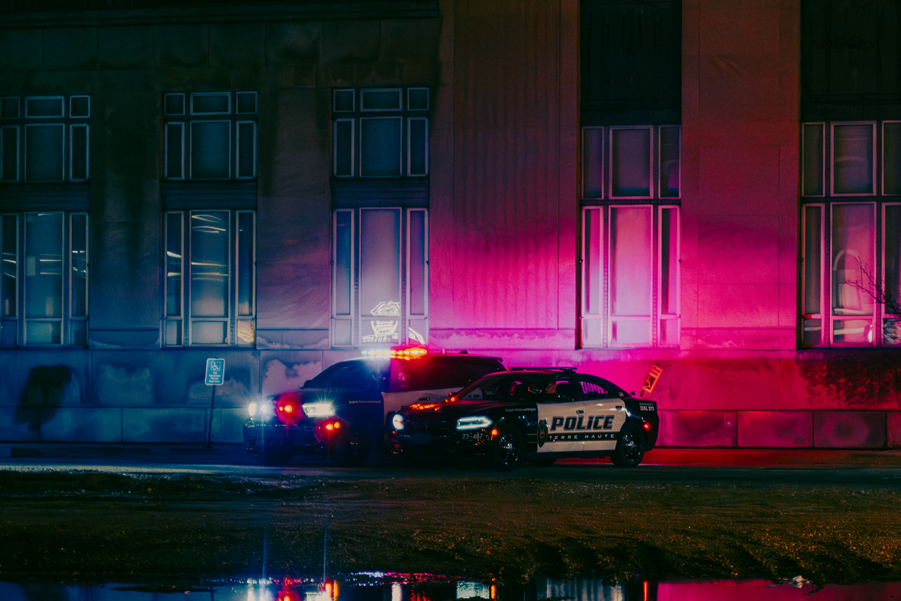 Two police cars parked in front of a building at night, with red and blue emergency lights flashing and casting colorful reflections on the wall.
