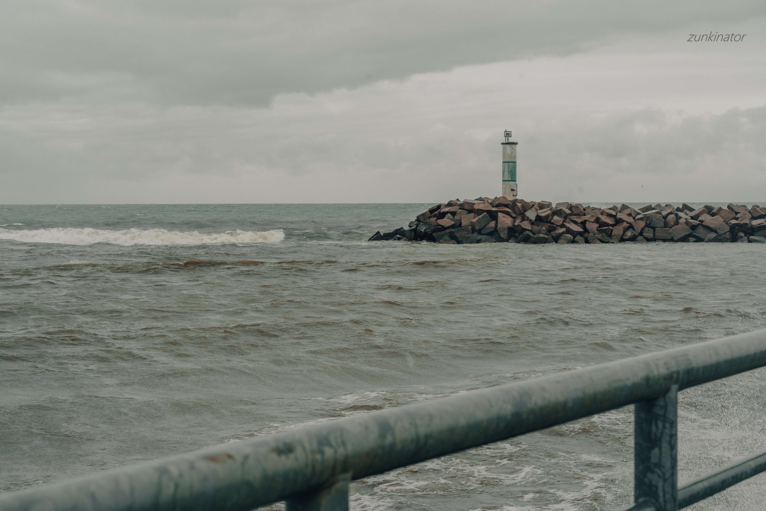 View of a breakwater with rocks and a lighthouse standing on it in the distance, seen from the shoreline with a metal railing in the foreground, under cloudy skies.