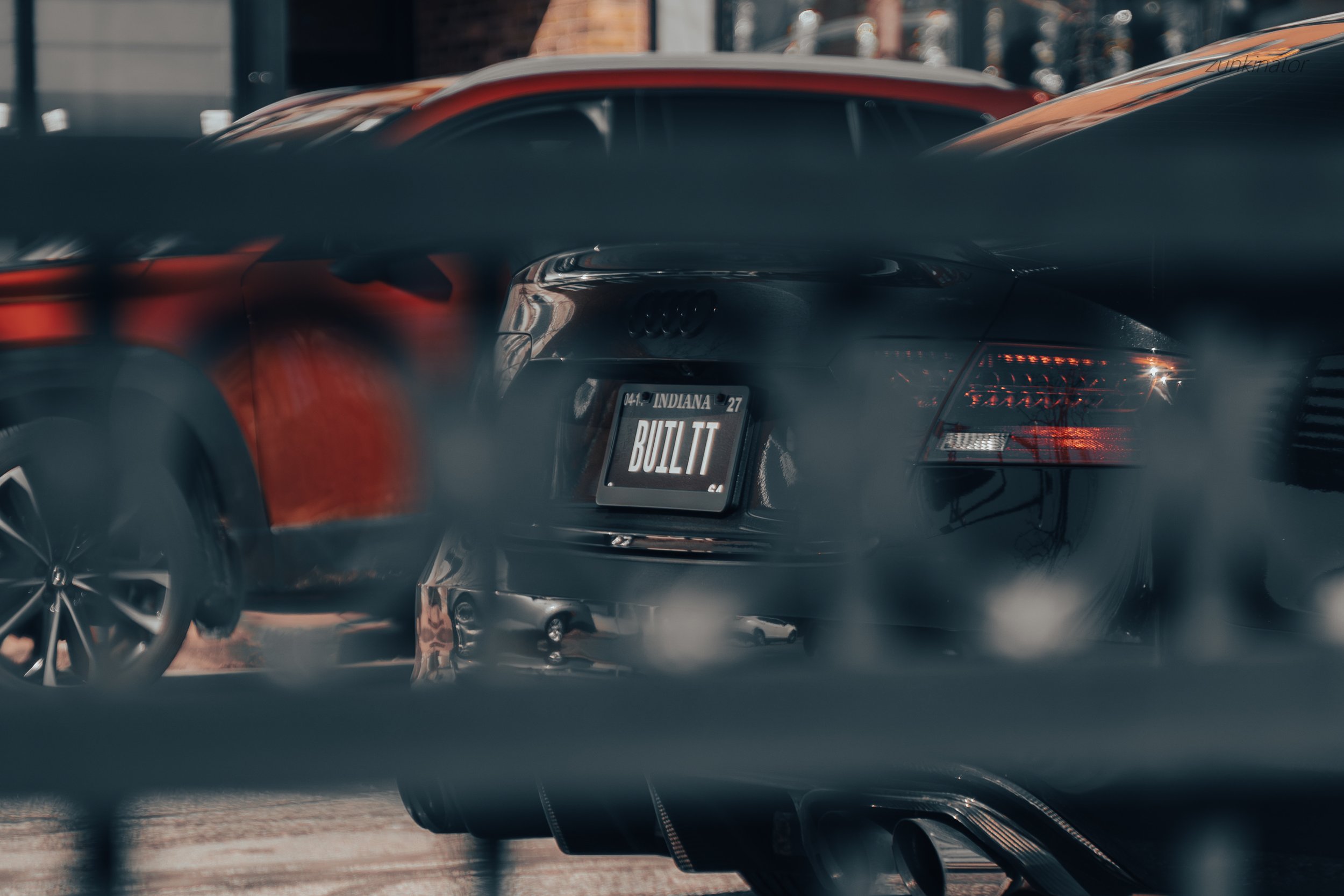 Black Audi car with a license plate that says 'BUILT T' parked behind a blurred metal fence, with other cars visible in the background.