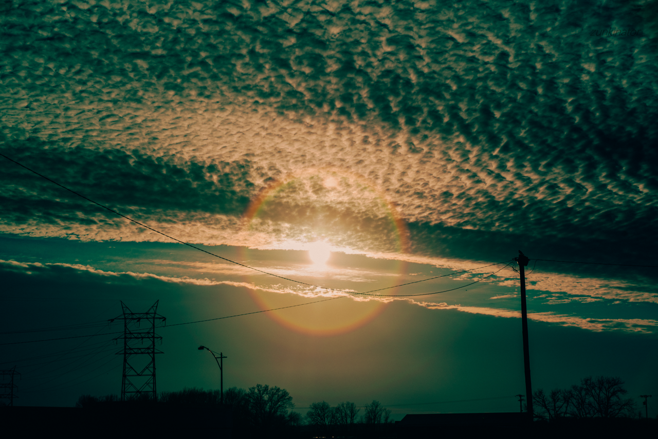 Sunset with a prominent sun halo, partly cloudy sky, power lines, utility poles, and silhouetted trees.