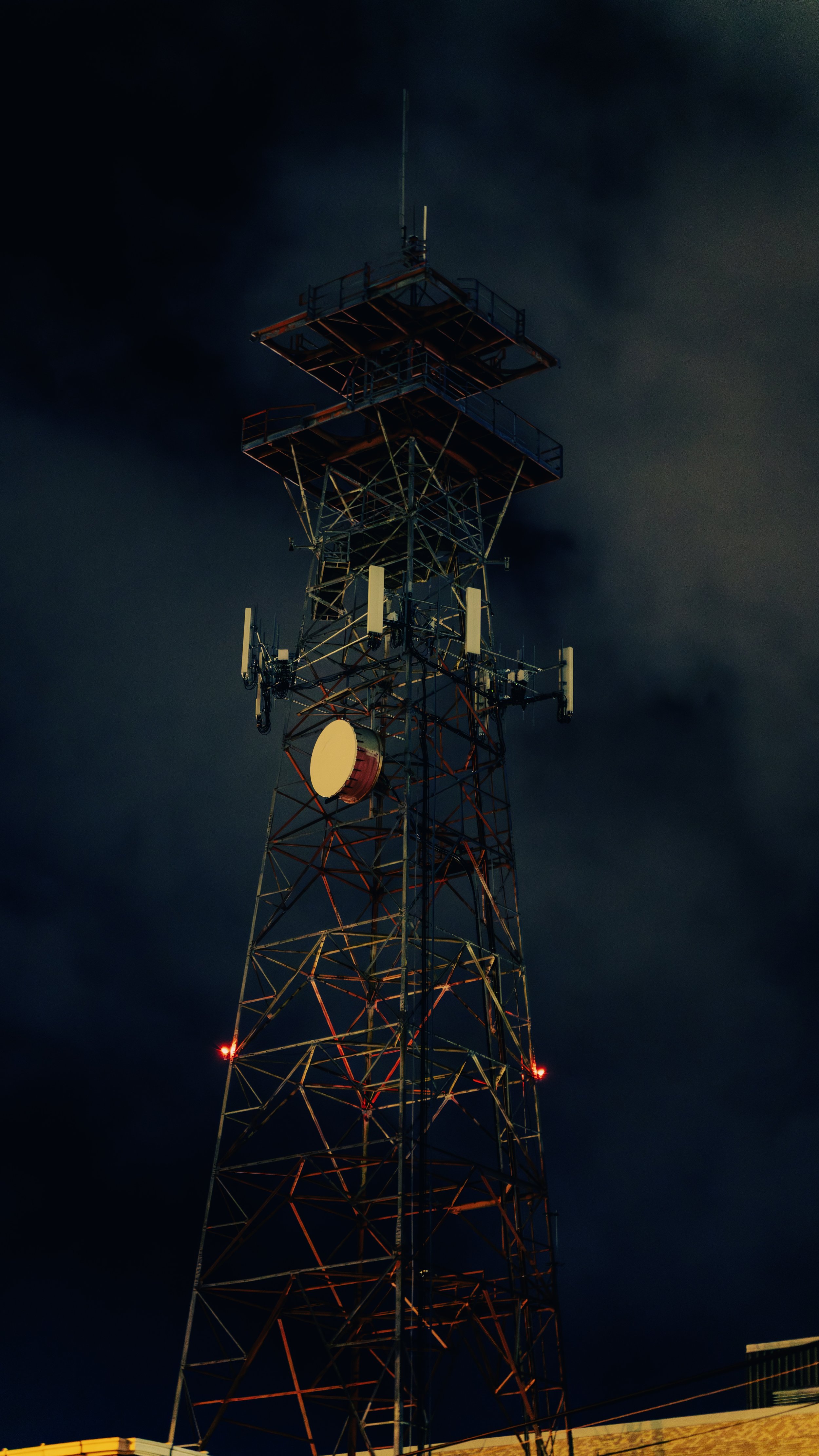 Nighttime view of a tall communication tower with multiple antennas and red warning lights, under a cloudy dark sky.