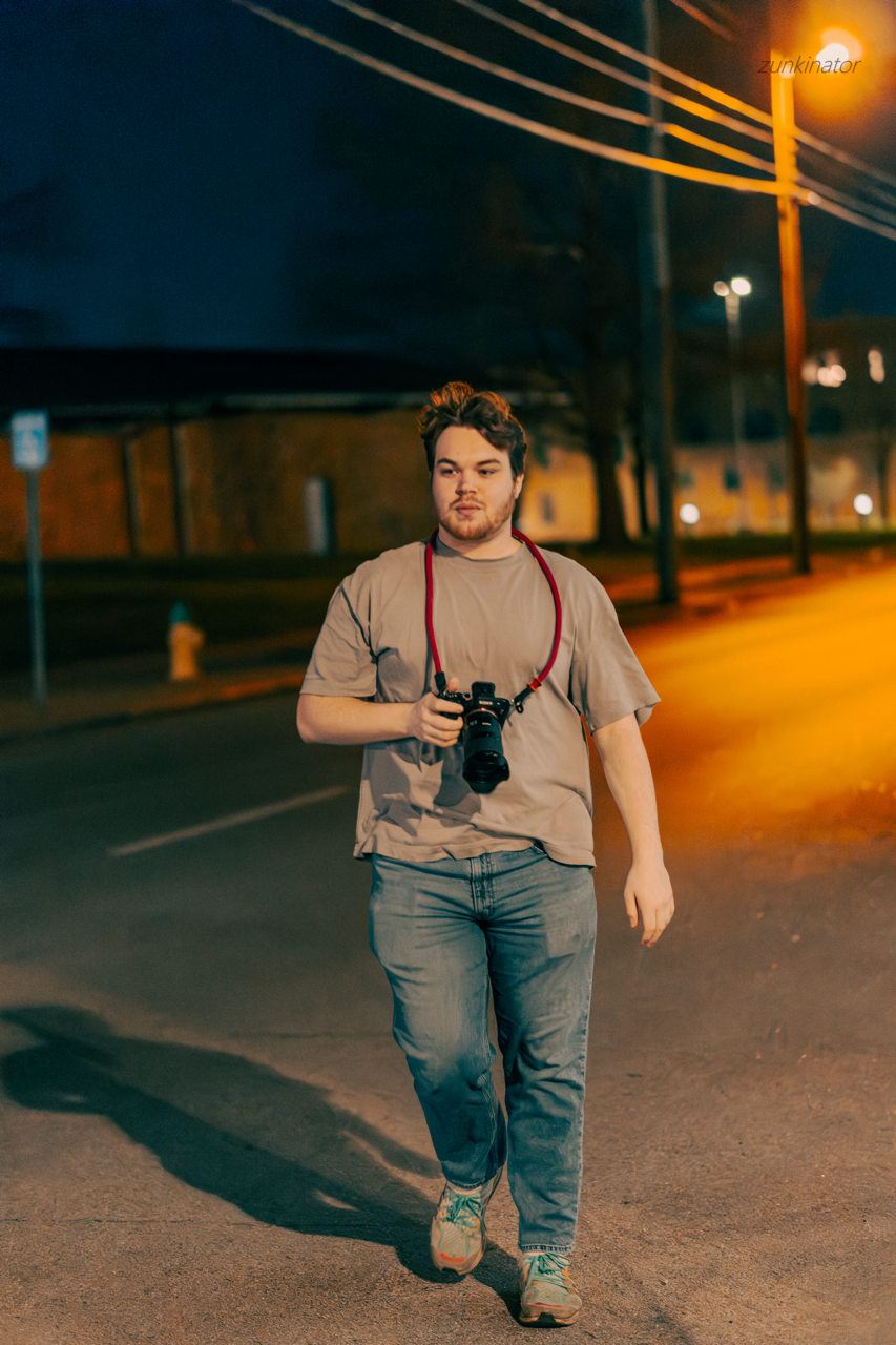 Young man walking on street at night with camera around his neck, dark background, streetlights, and power lines.