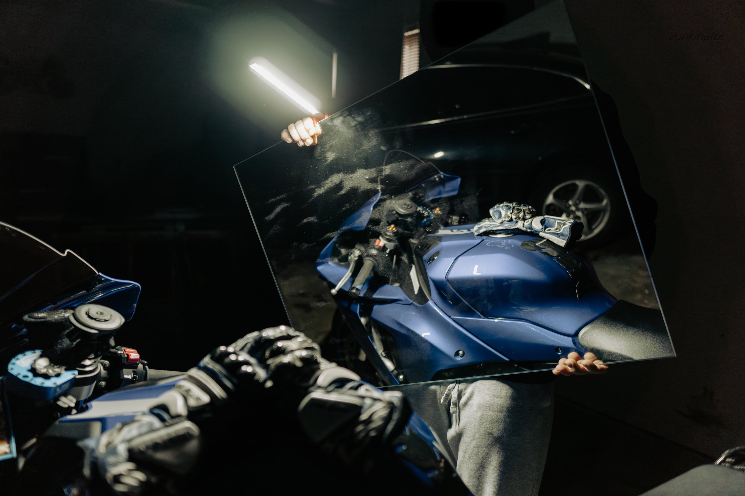 Person holding a mirror reflecting a blue motorcycle with gloves on the seat and a car in the background.