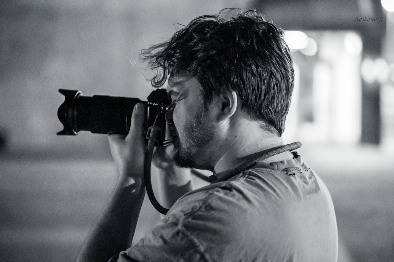 A man with wavy hair and a beard is holding a camera up to his eye, taking a photo in an outdoor setting at night.