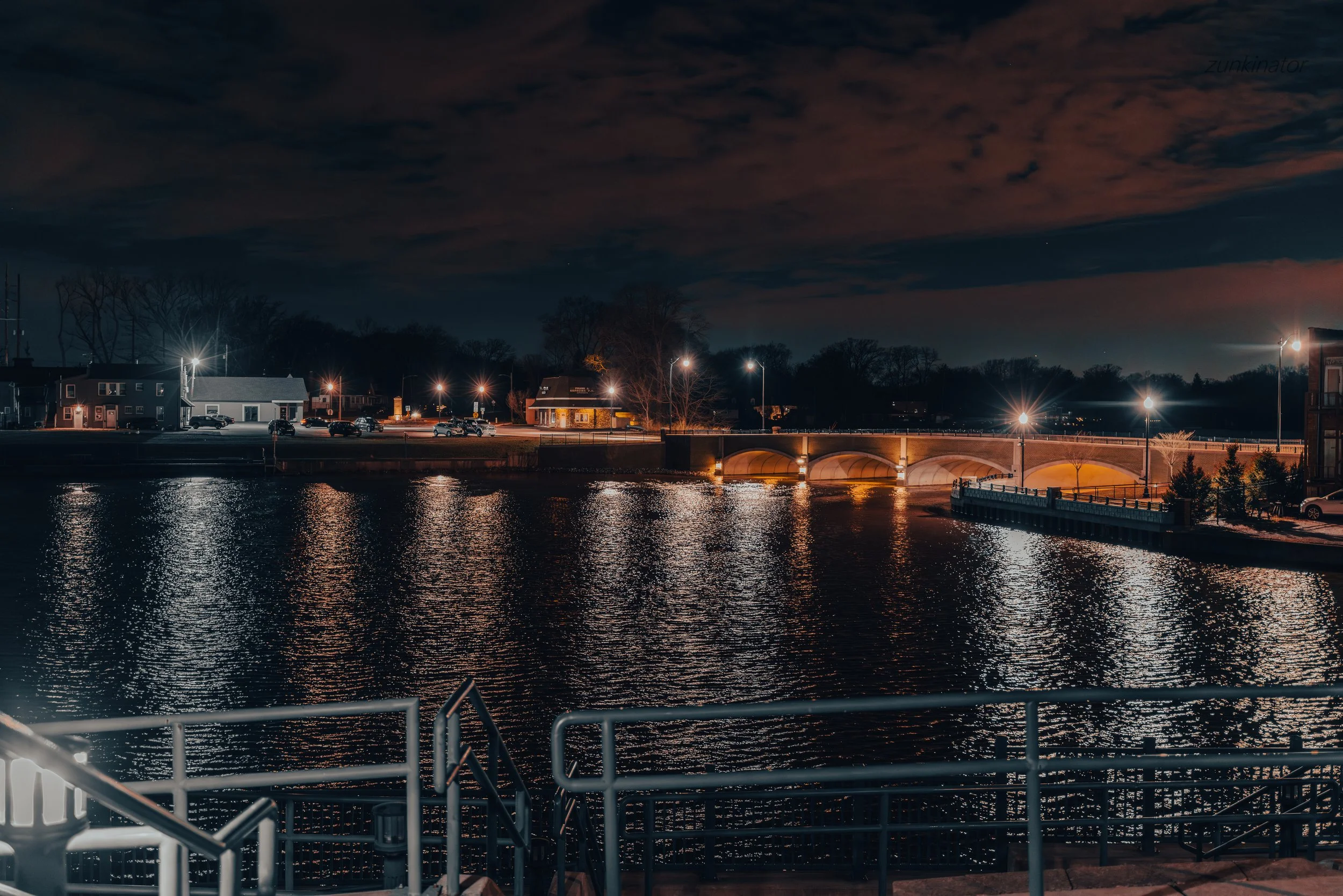 Nighttime view of a river with reflections of streetlights, a bridge illuminated with warm lights, and buildings with lit windows in the background.