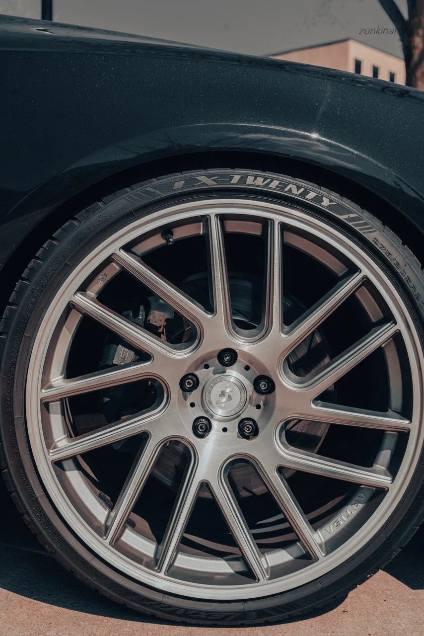 Close-up of a car wheel with a multi-spoke alloy rim and tire, showing the tire's sidewall details and part of the car body.
