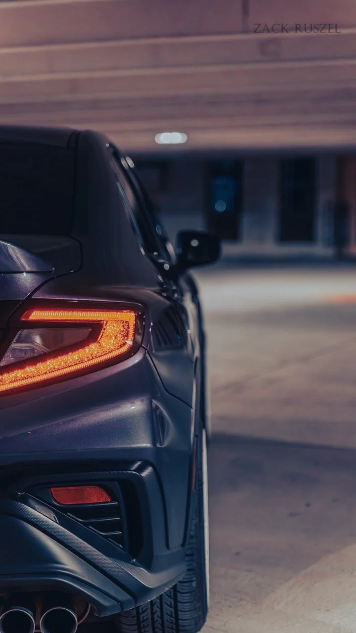 A close-up view of the rear left side of a black sports car parked indoors, showing the tail light, part of the rear bumper, and tire, with a wooden ceiling and a blurred background.