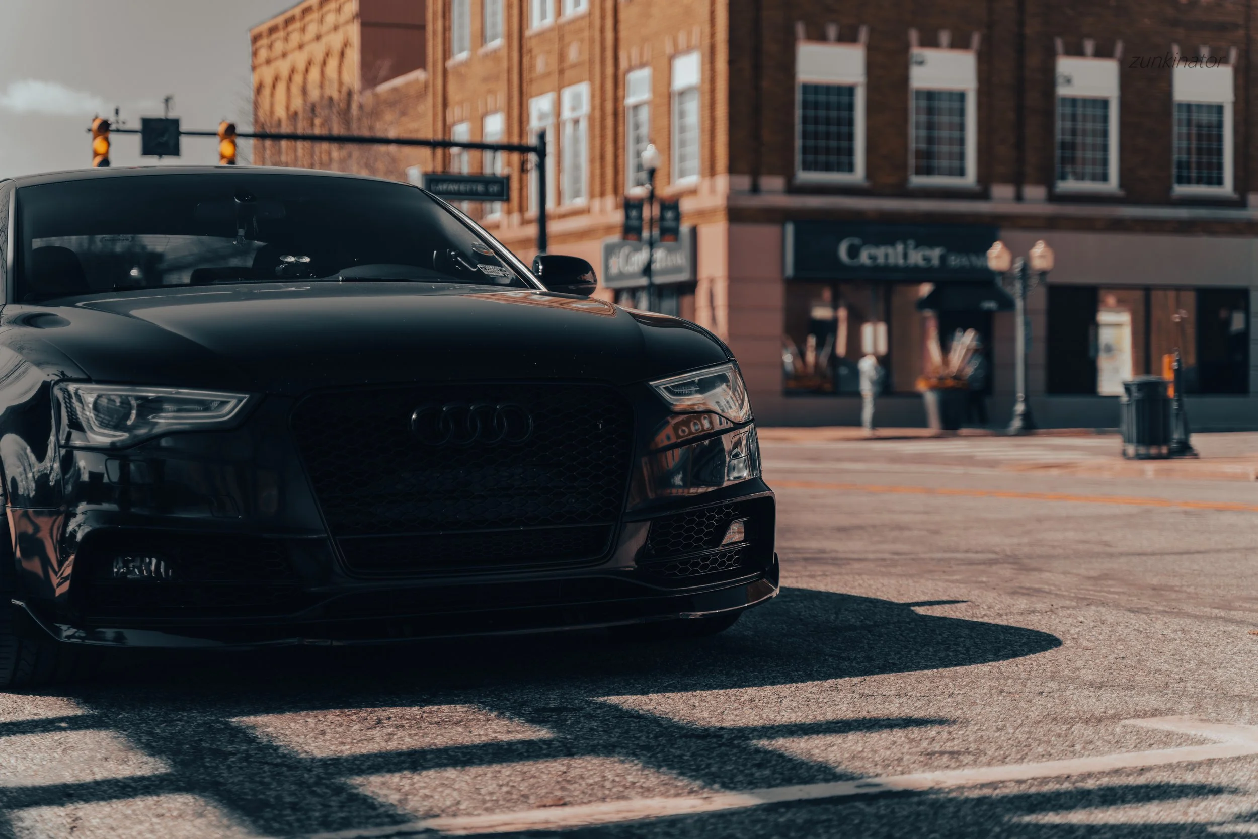 A black Audi car parked on an urban street with brick buildings, a storefront named 'Center' in the background, and a traffic light overhead. The car is partially in shadow, reflecting nearby structures.