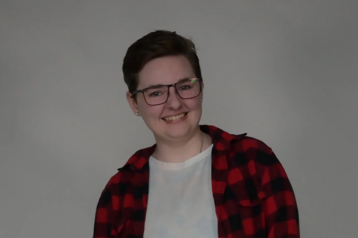 A young person with short brown hair, glasses, and earrings, smiling while wearing a white shirt and red plaid jacket, standing against a gray background.