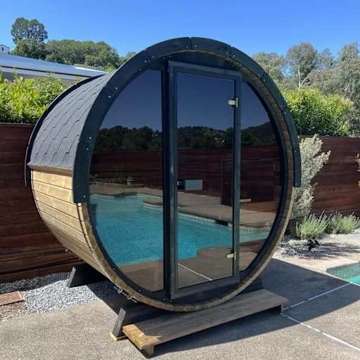 A small, round wooden sauna with a glass door, situated on a concrete patio beside a swimming pool, surrounded by a wooden privacy fence and greenery.